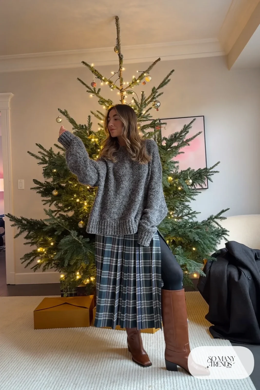 Woman in an oversized gray sweater, slate and wine plaid pleated midi skirt, sheer tights and tall brown boots by a tree, outfit 10