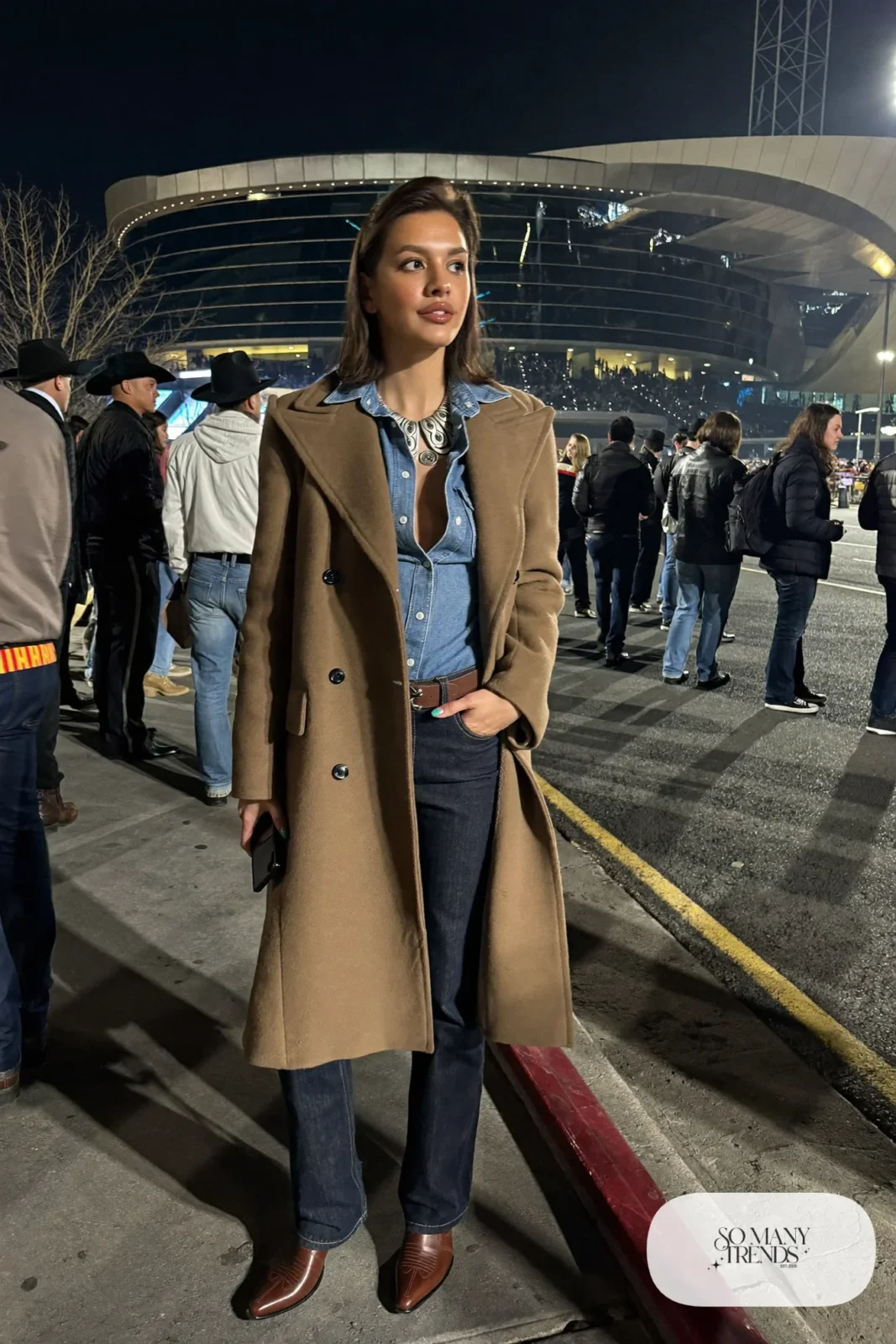 Woman in long camel wool coat, chambray western shirt, dark straight jeans and brown boots, NFR outfit 12