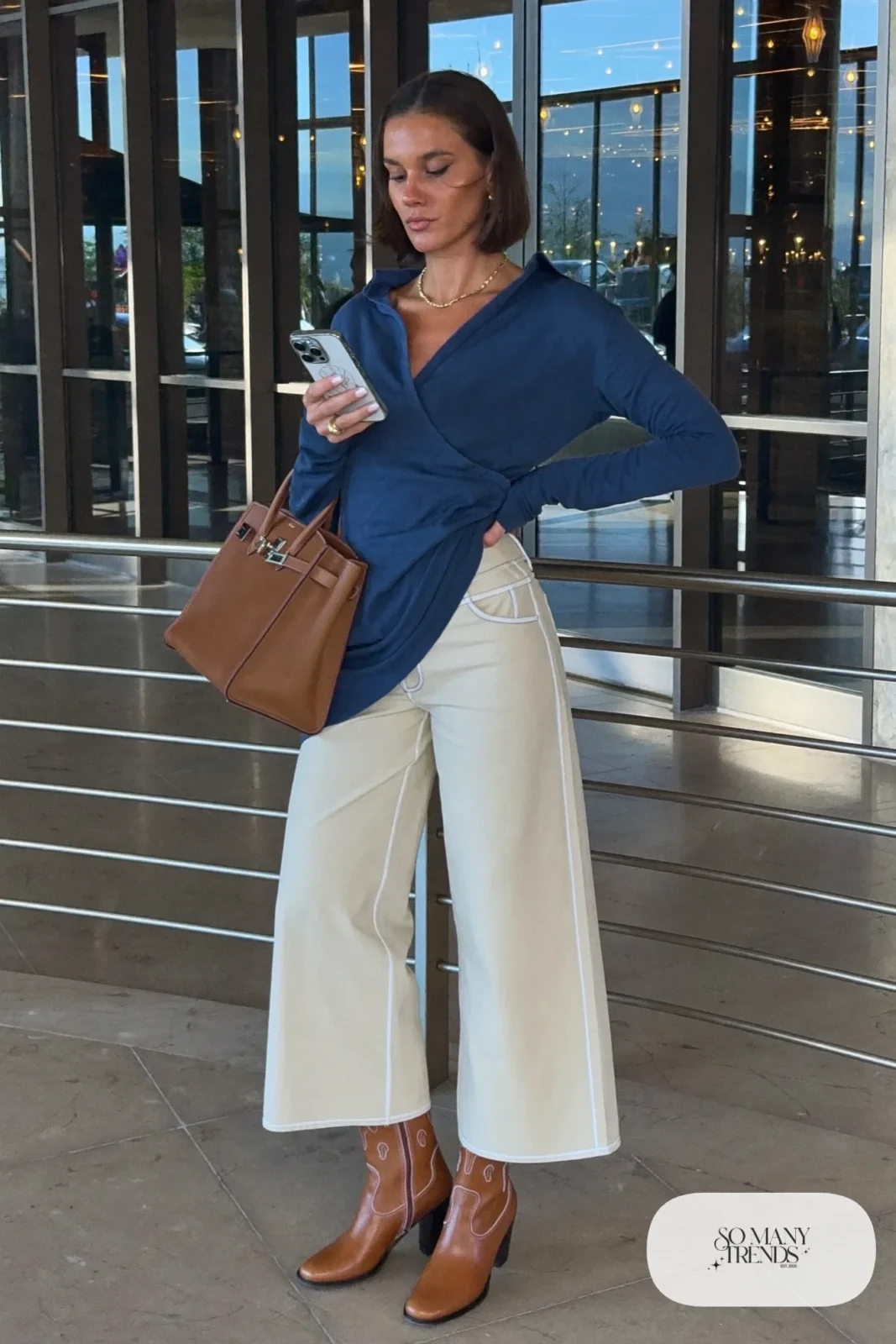 Woman in ink blue wrap top, cream wide-leg pants, cognac boots and leather tote, NFR outfit 19