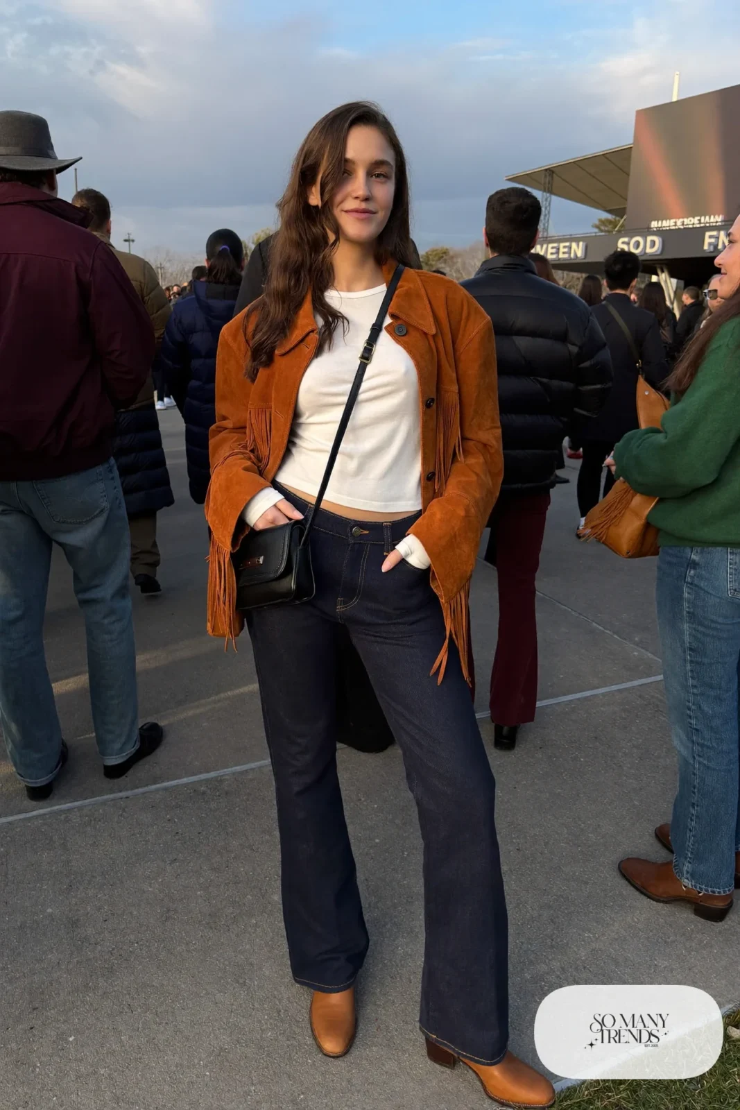 Woman in a cognac suede fringe jacket, white ribbed top, navy flared jeans and tan western boots, concert outfit 1