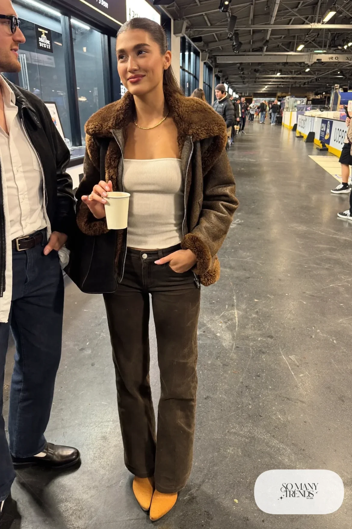 Woman in brown faux shearling jacket, cream ribbed knit top, dark straight jeans and tan suede boots, hockey game outfit 3