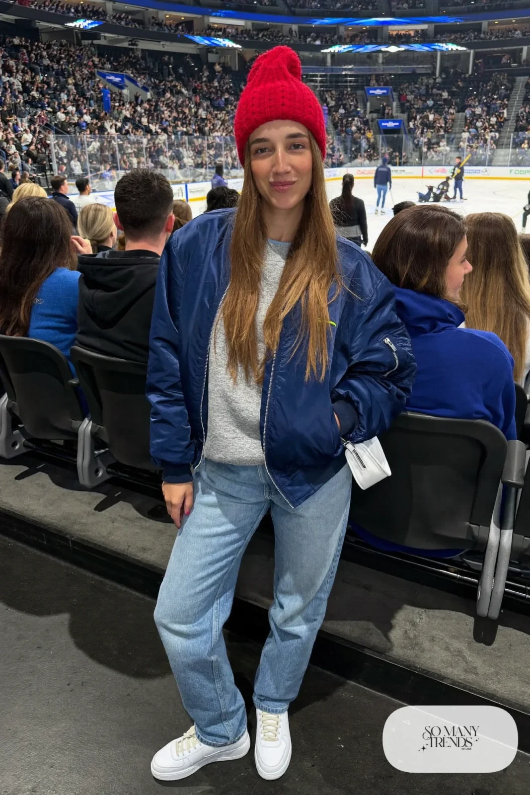 Woman in navy bomber jacket, heather gray sweatshirt, light jeans, red knit beanie and white sneakers, hockey game outfit 6