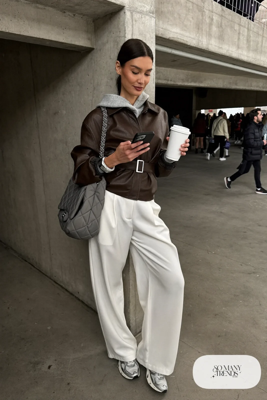Woman in brown faux leather jacket, heather gray hoodie, white pleated wide-leg pants and chunky gray sneakers, hockey game outfit 7