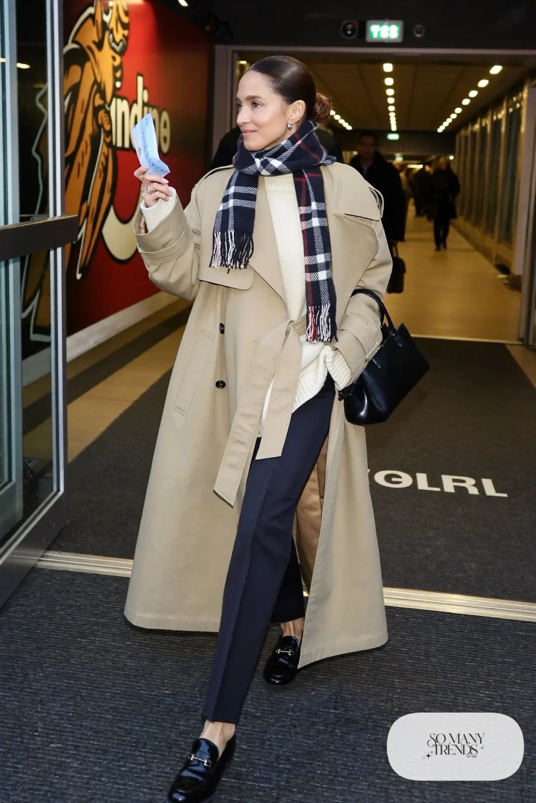 Woman in beige lined trench coat, chunky cream sweater, dark straight pants, plaid scarf and black loafers, hockey game outfit 14