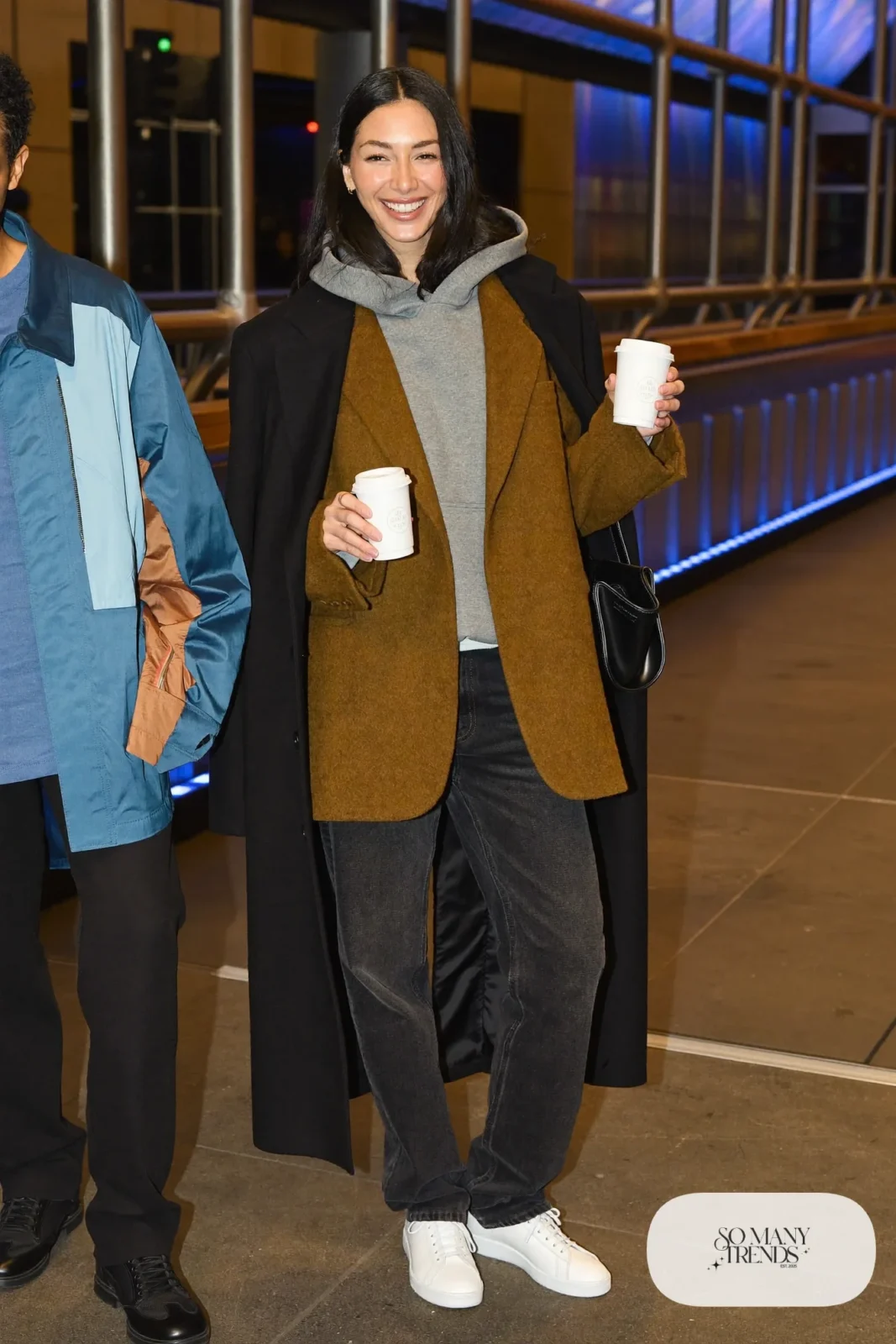 Woman in camel oversized blazer, heather gray hoodie, dark straight jeans, long black coat and white sneakers, hockey game outfit 19
