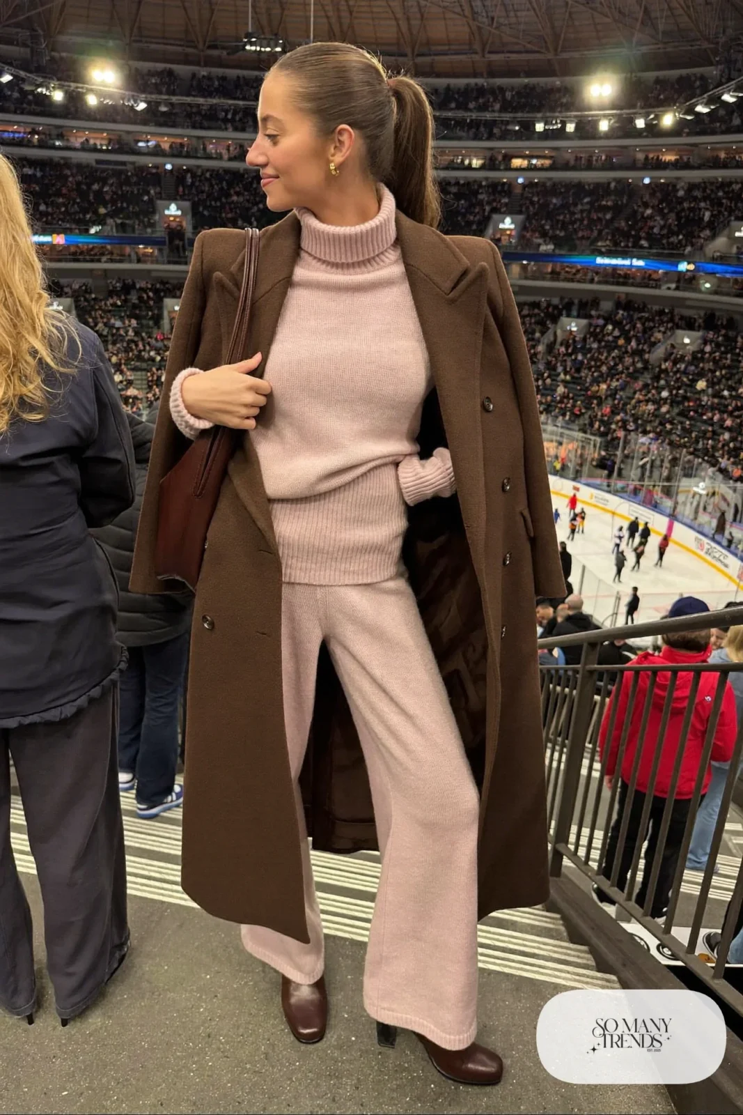 Woman in pale pink knit sweater and pants set, long espresso coat, brown boots and brown shoulder bag, hockey game outfit 24