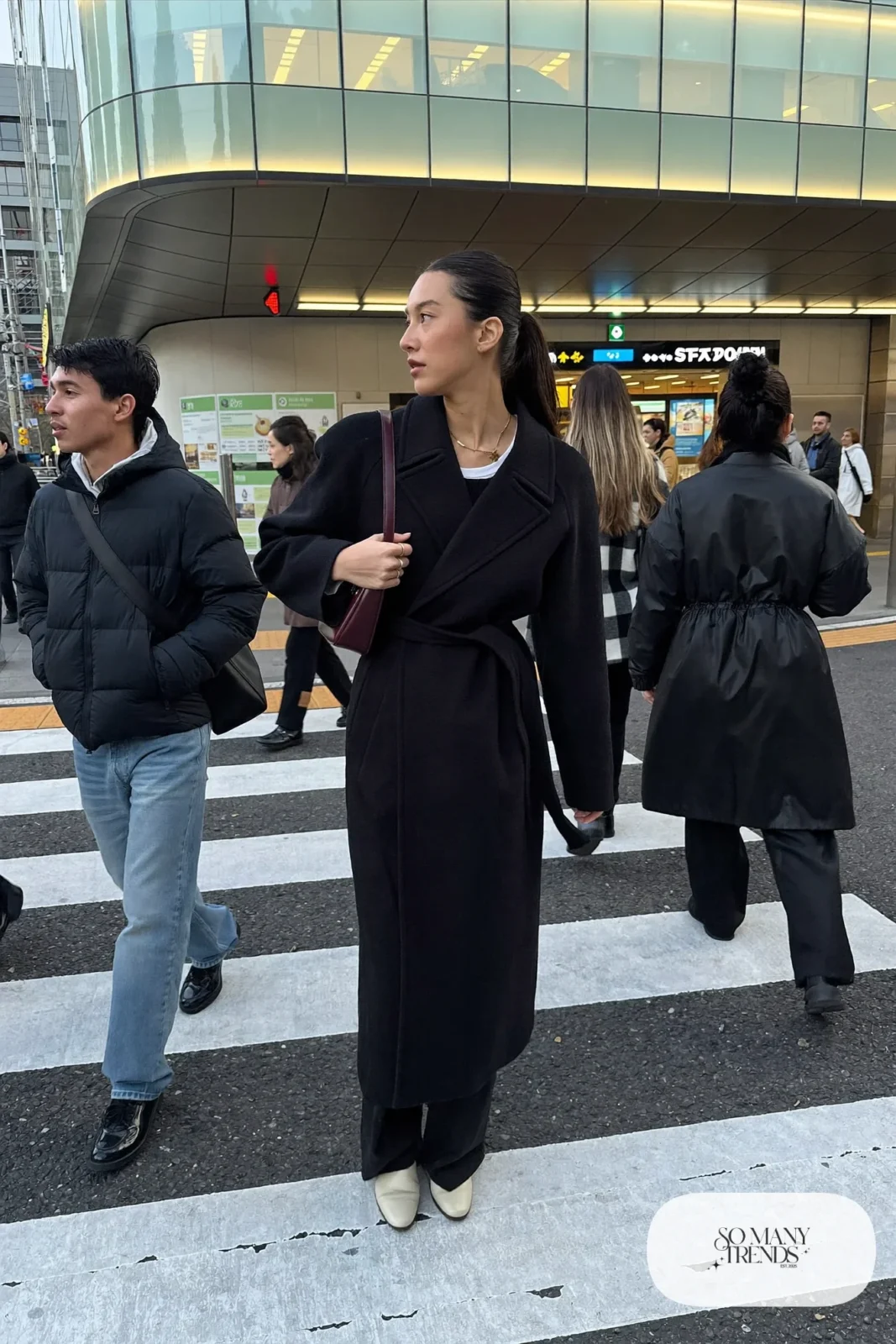 Woman in a black wool wrap coat with a tied sash, black wide-leg pants, cream flat ankle boots and a burgundy bag, outfit 2