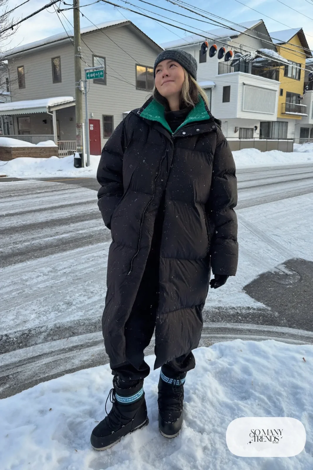 Woman in a long black hooded puffer coat, emerald knit sweater, black snow pants and waterproof snow boots in deep snow, outfit 5