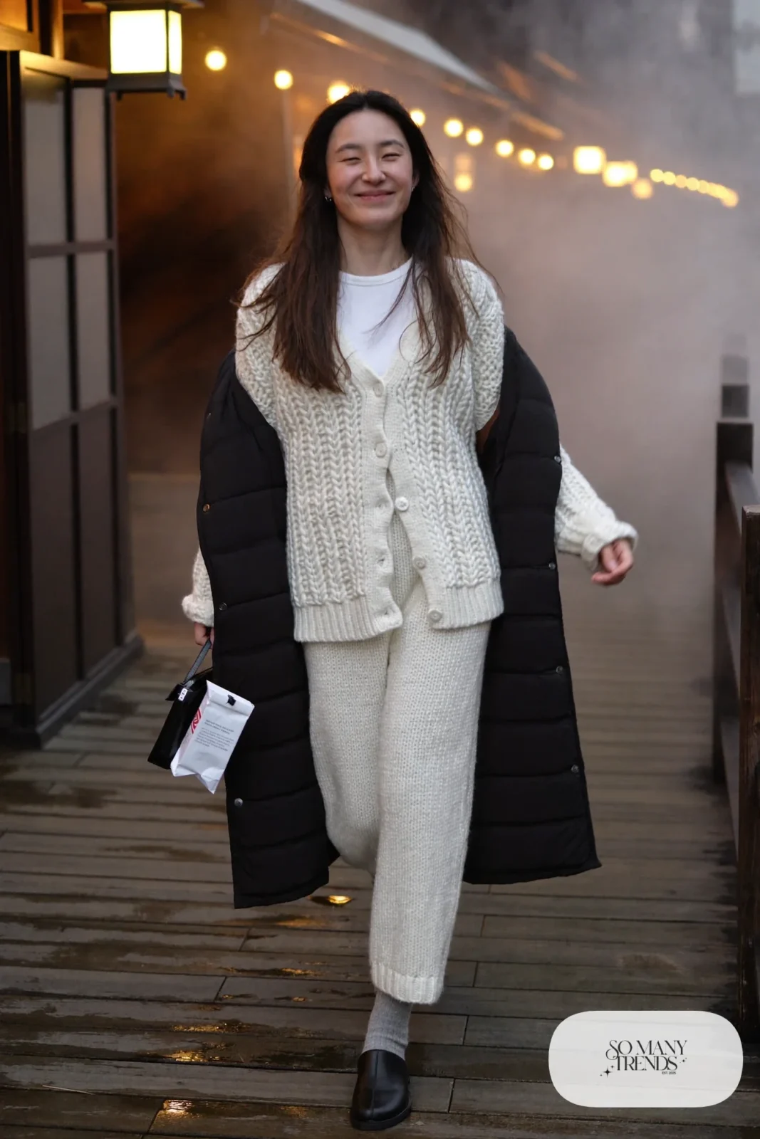 Woman in a pearl knit cardigan and wide pants set, black quilted coat over her arm and leather slip-on shoes in an onsen town, outfit 6