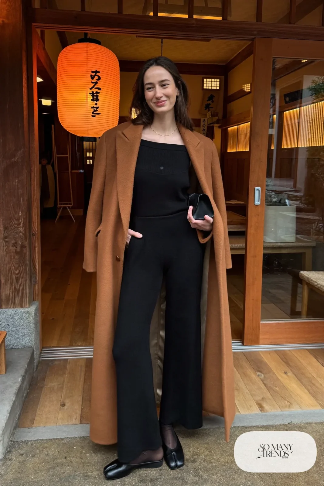 Woman in a black wide-leg knit jumpsuit with a camel wool coat and black leather slip-on flats at a ryokan entrance, outfit 22