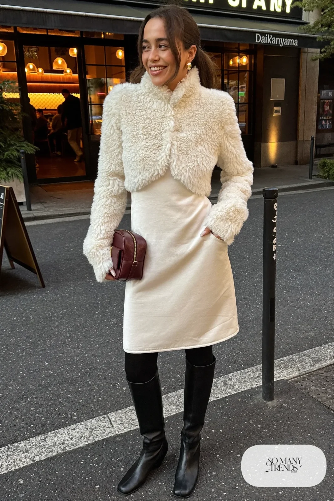 Woman in a cropped shearling jacket over a pearl midi dress with tall black boots and a burgundy bag in Tokyo, outfit 24