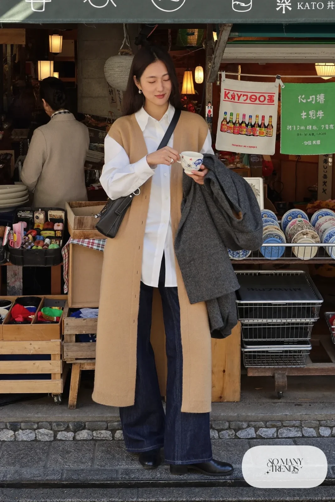 Woman in a camel long knit vest over a white shirt, wide-leg dark jeans, black boots and a gray coat over her arm, outfit 27