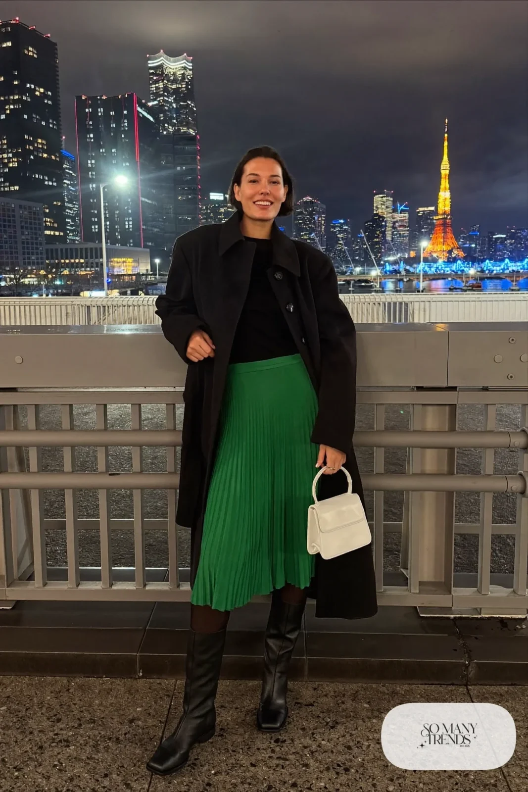 Woman in a long black coat over a fitted black knit top, emerald pleated midi skirt and tall black boots with a pearl bag in Tokyo at night, outfit 31