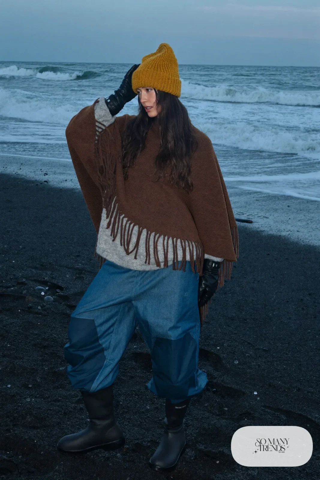 Woman in a brown wool poncho, denim blue insulated snow pants, black waterproof boots and mustard knit hat, outfit 3