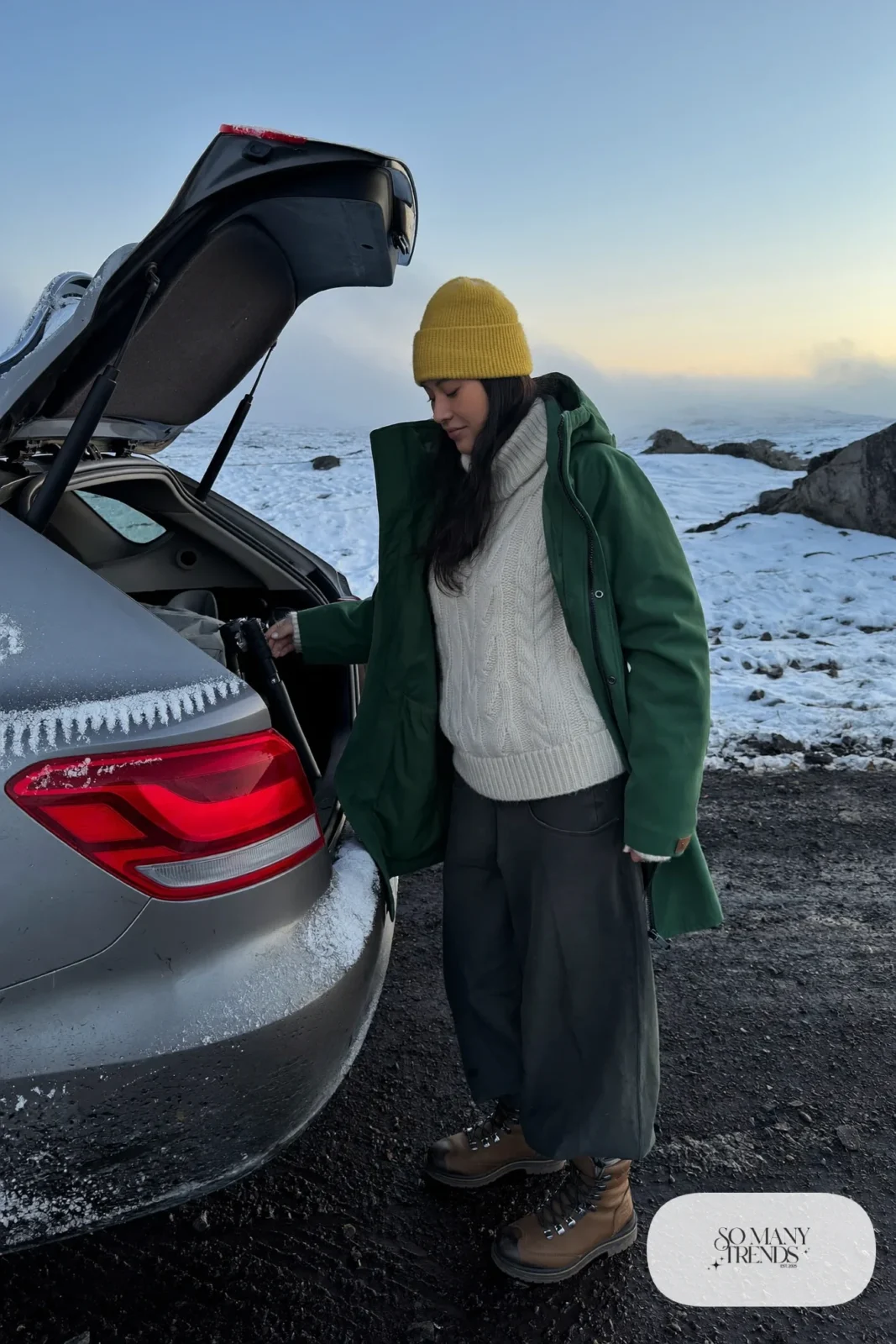 Woman in a forest green waterproof shell, cream cable-knit sweater, dark wide pants and waterproof boots on a snowy road, outfit 5