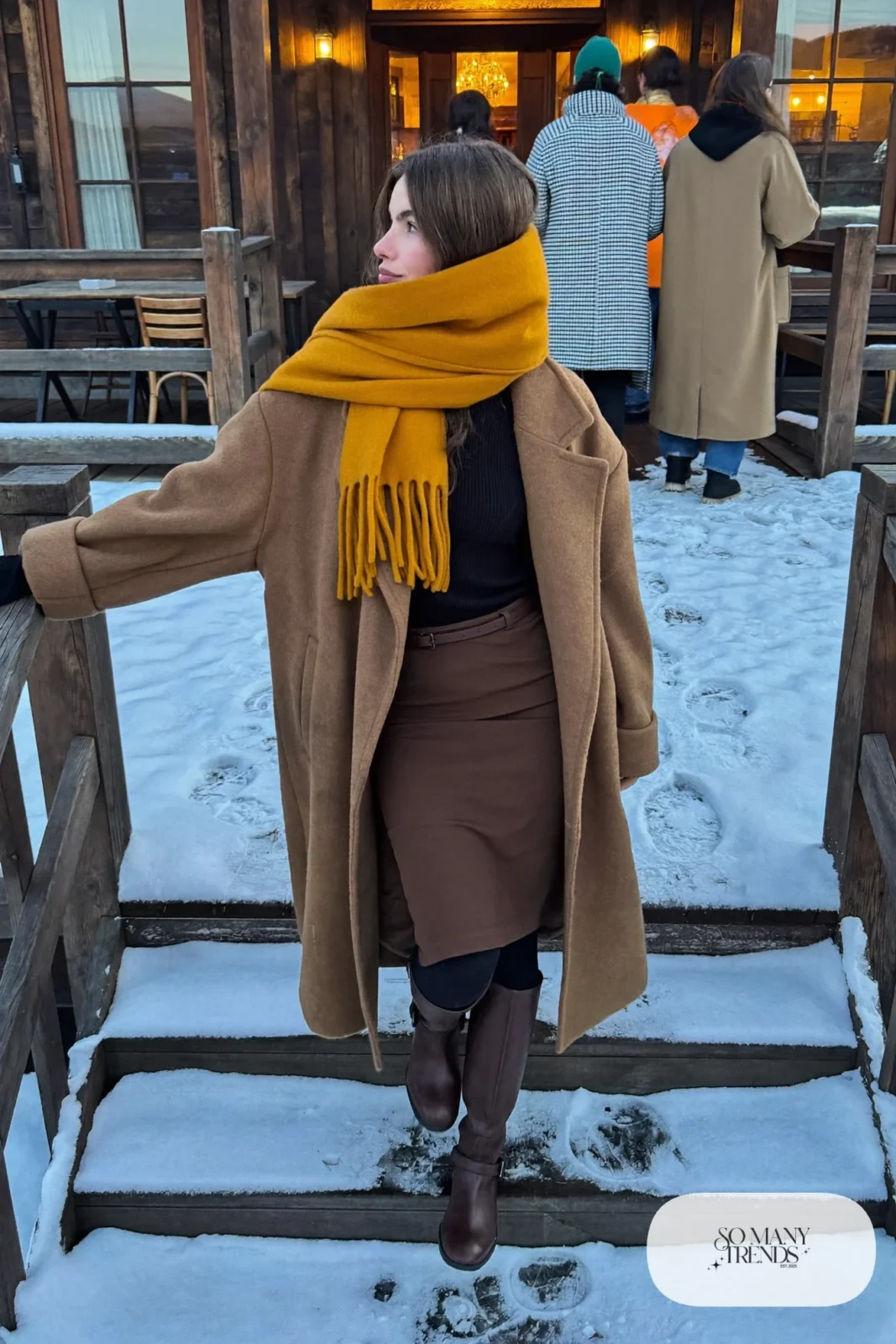 Woman in a long camel coat, chocolate wool midi skirt, black tights, tall brown boots and a mustard scarf, outfit 6