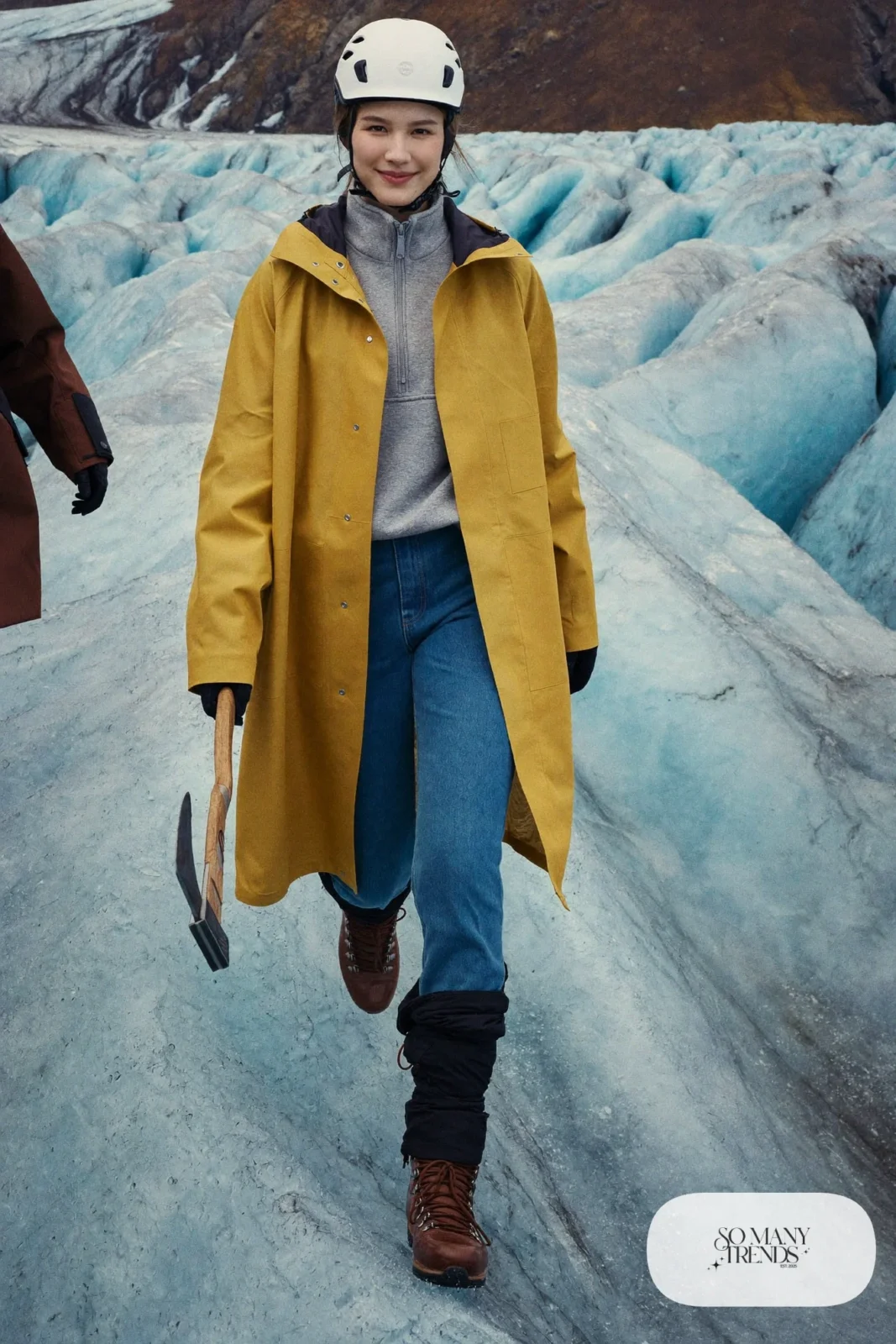 Woman in a mustard waterproof shell, denim blue stretch hiking pants, hiking boots, helmet and gloves on a glacier, outfit 7