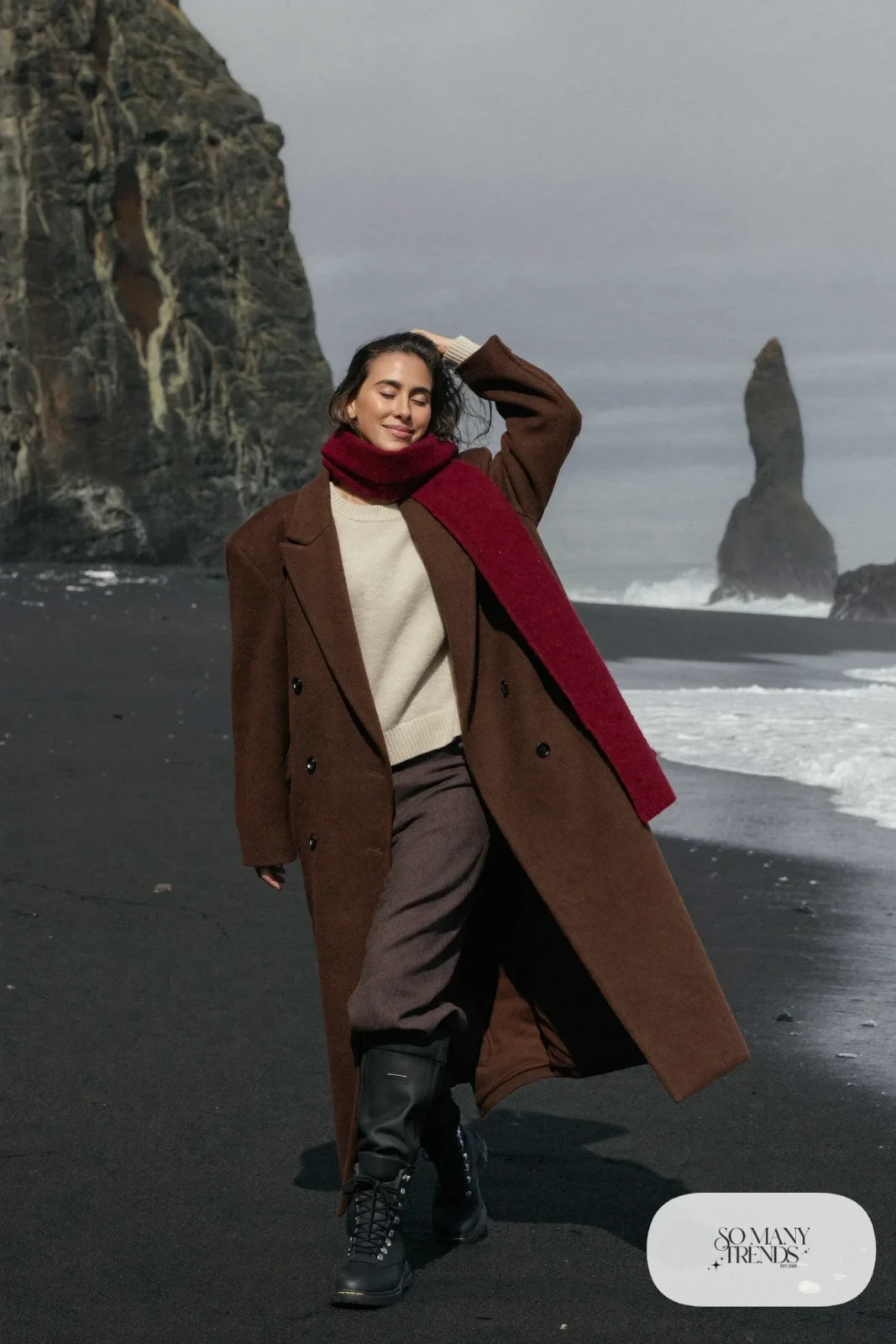 Woman in a long chocolate wool coat, cream knit sweater, wide gray pants and waterproof boots on black sand beach, outfit 9