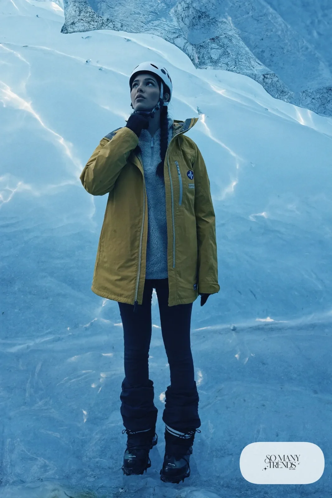 Woman in a mustard shell, gray fleece, black thermal pants and boots inside a blue ice cave, outfit 13