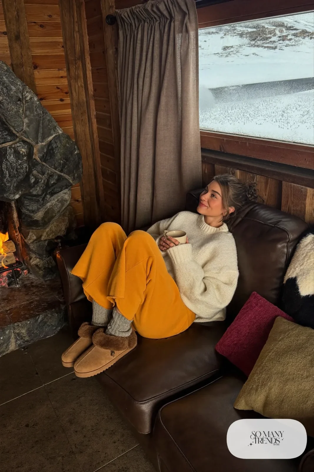 Woman in a cream chunky knit sweater, mustard wide knit pants and shearling slippers by a lodge fireplace, outfit 16