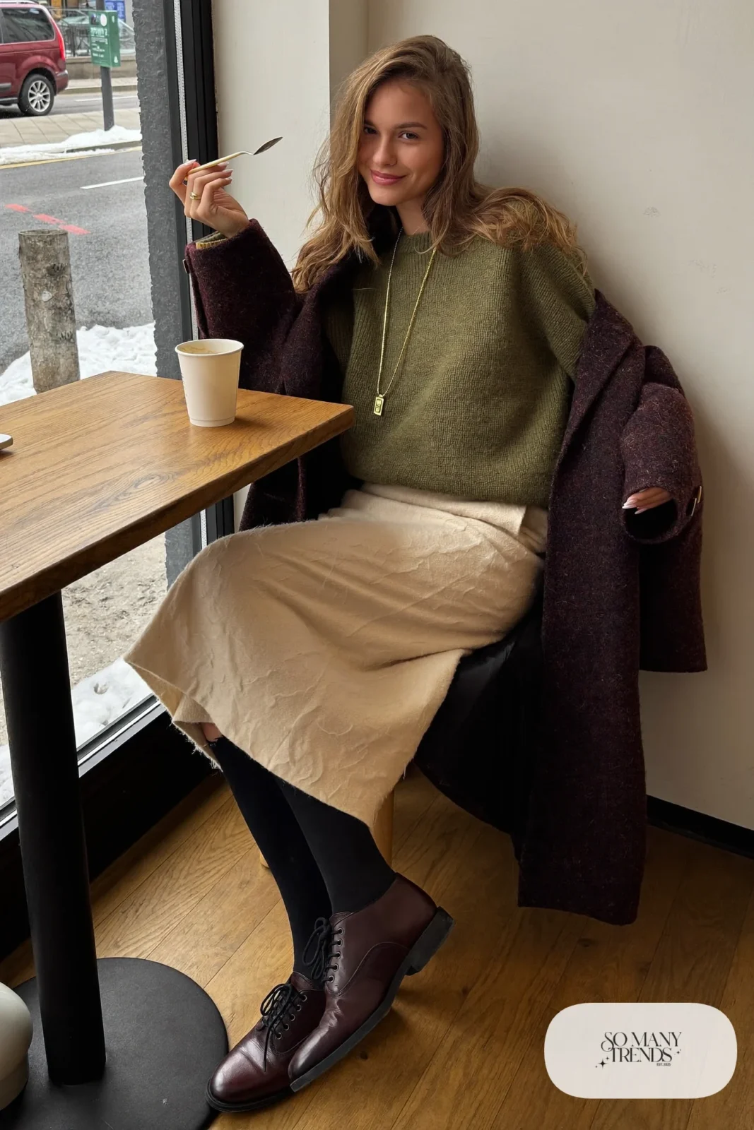 Woman in an olive boxy sweater, cream pencil skirt, black tights, burgundy lace-up boots and a long coat in a museum cafe, outfit 22
