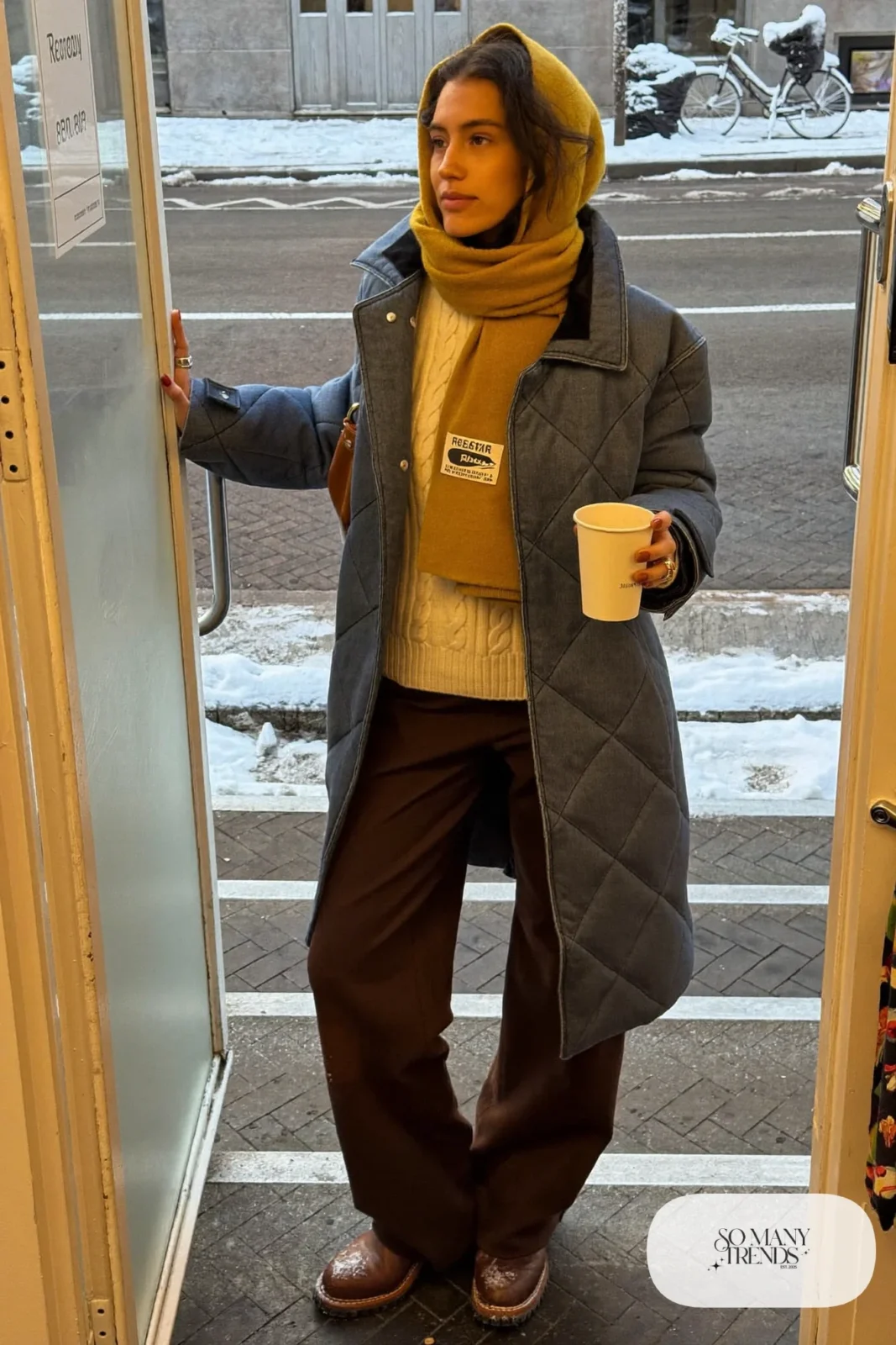Woman in a denim blue belted quilted coat, cream knit, brown boots and wool scarf in a Reykjavik cafe doorway, outfit 24