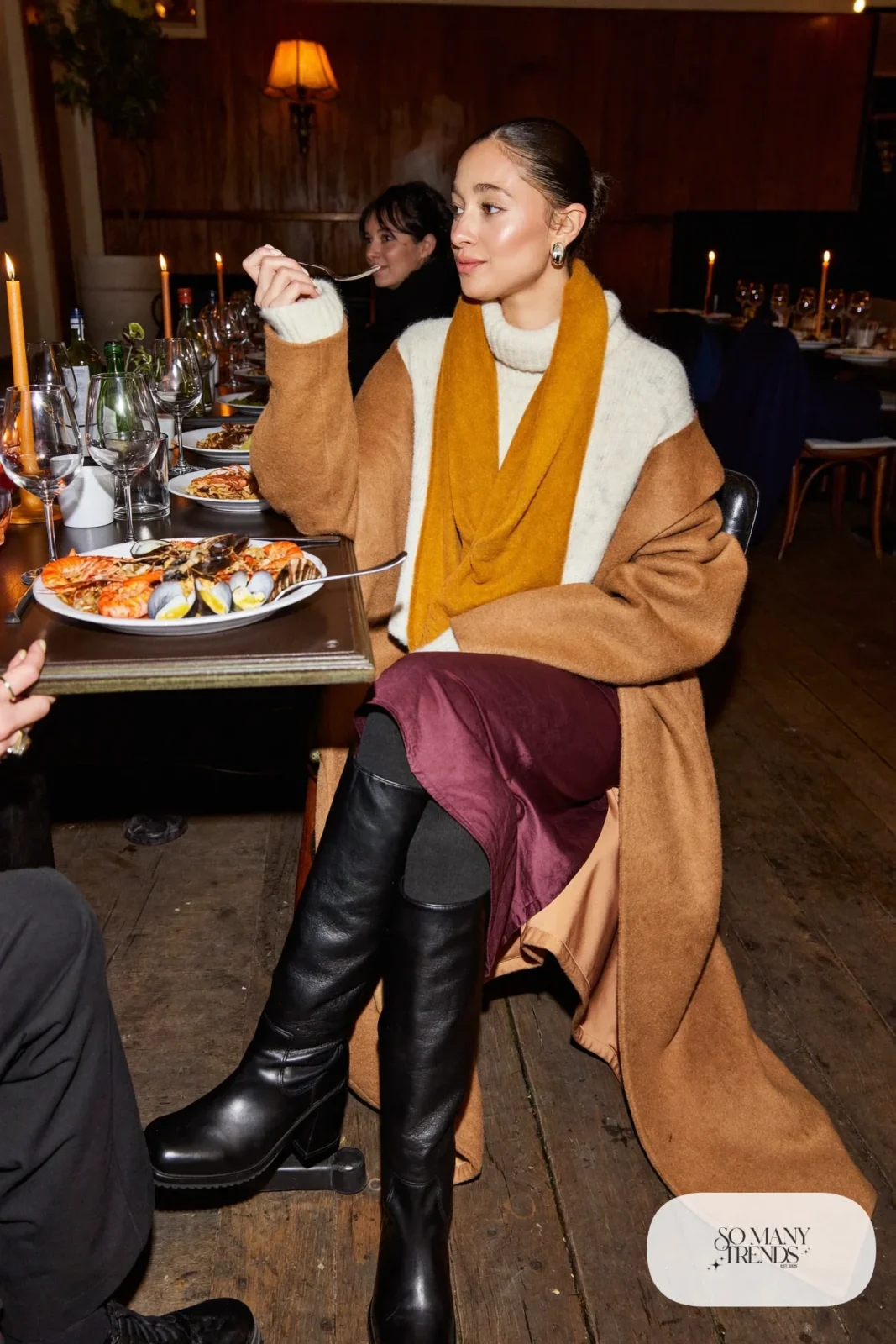 Woman in a burgundy satin midi skirt, cream chunky knit, tall black boots and a camel coat leaving a Reykjavik restaurant, outfit 27