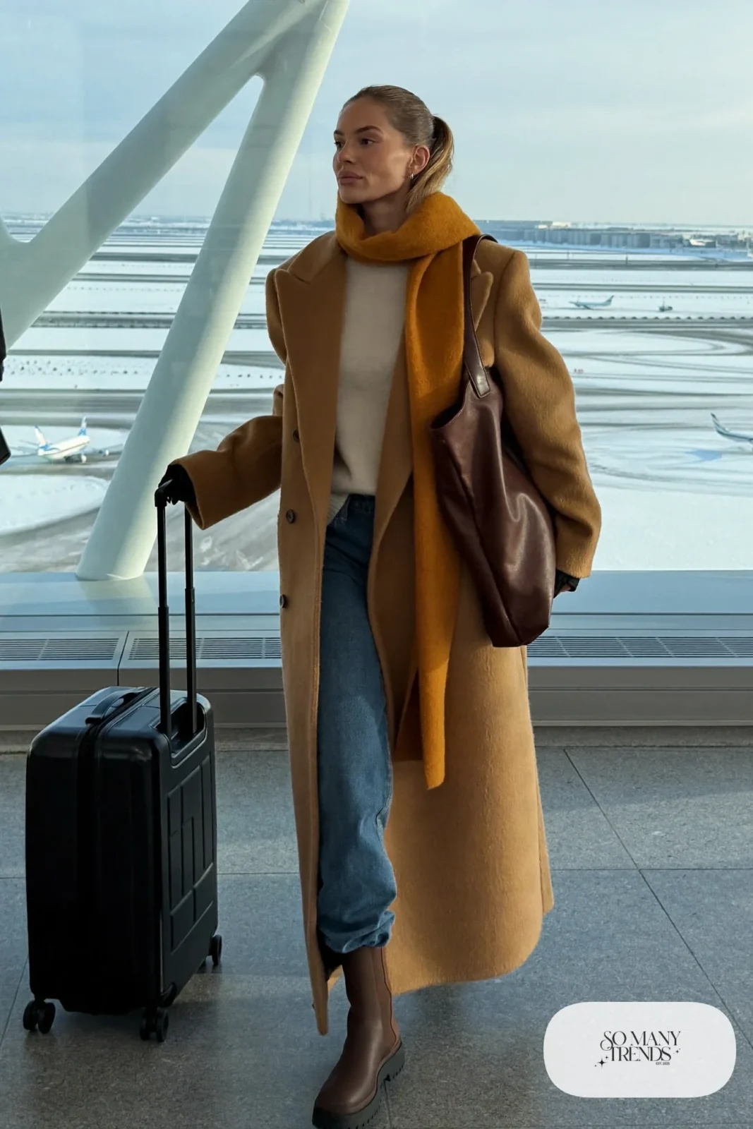 Woman in a long camel coat, cream sweater, blue jeans, brown boots and a big scarf at Keflavik airport, outfit 30