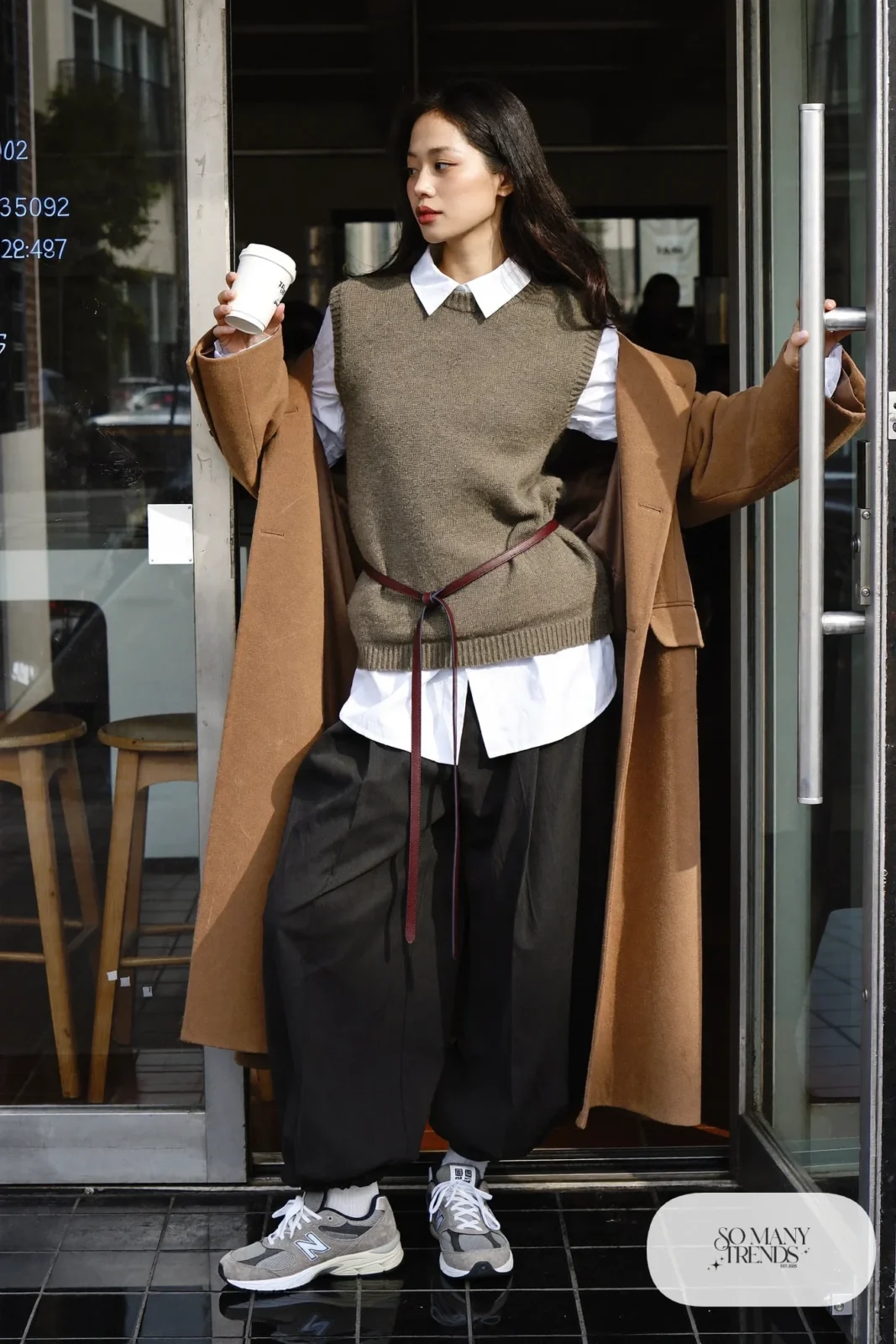 Woman in taupe knit vest, white shirt, thin low belt, wide black pants and gray New Balance 9060 sneakers, outfit 10