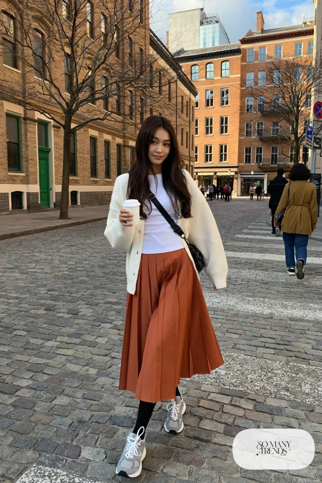 Woman in terracotta pleated midi skirt, cream chunky cardigan, black tights and gray New Balance 9060 sneakers, outfit 11