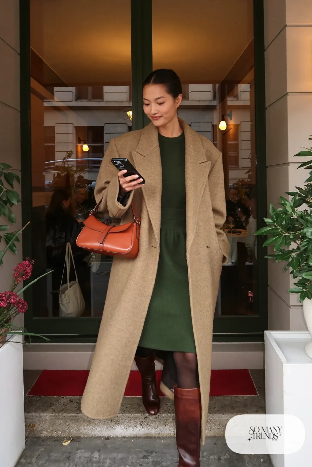 Woman in long camel coat, forest green midi dress, sheer tights and tall brown boots, outfit 2