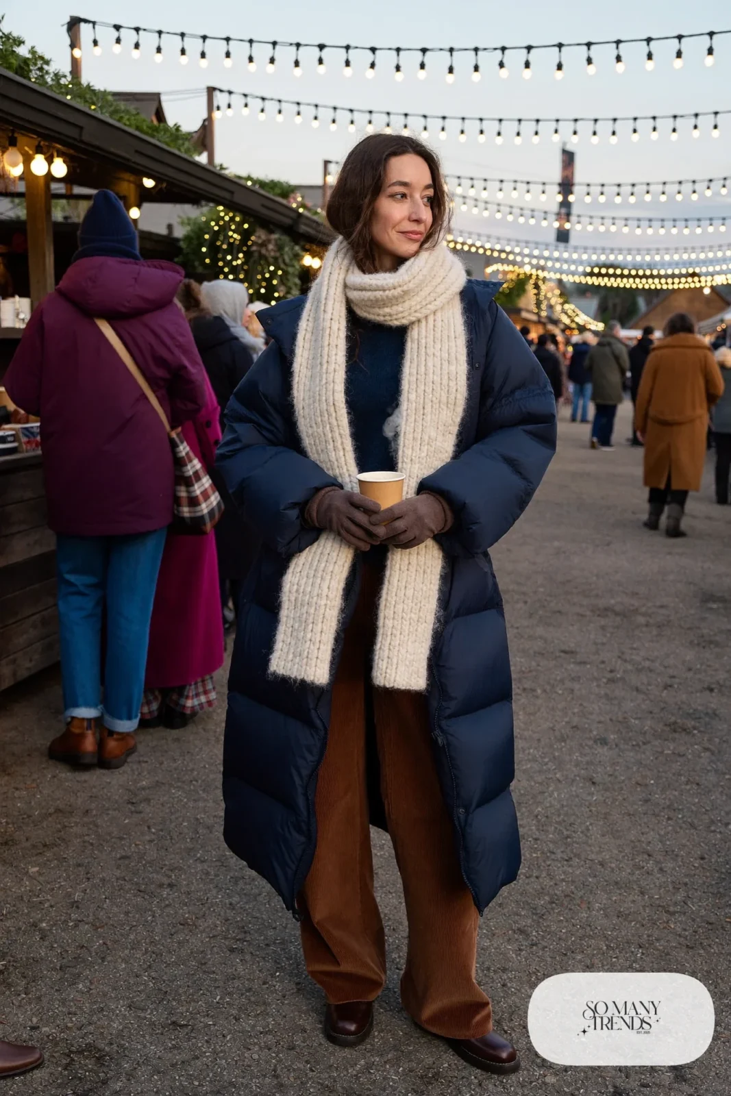 Woman in a long navy puffer coat, brown corduroy pants, cream knit scarf and tobacco leather boots, outfit 9