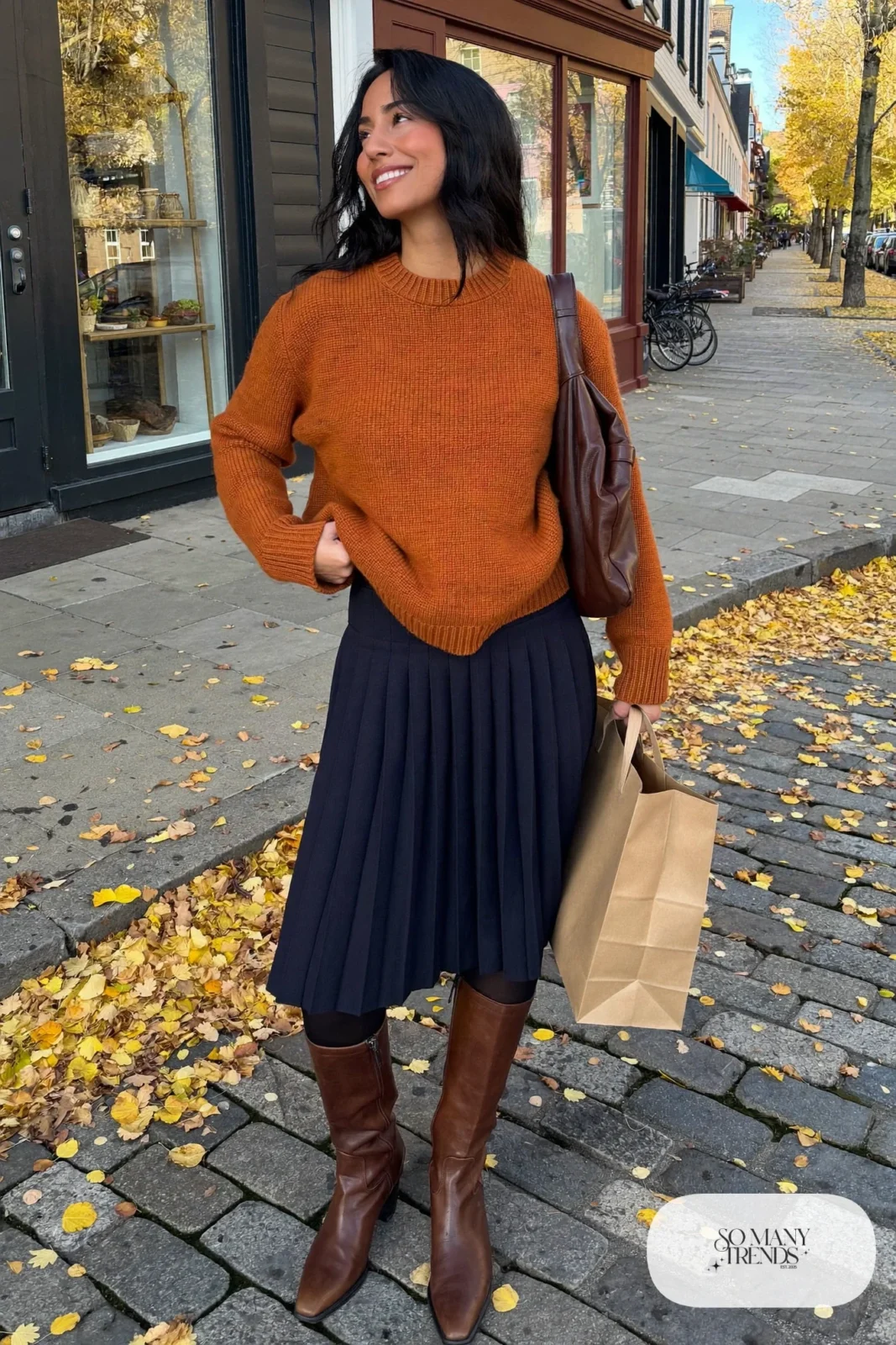 Woman in a rust sweater, navy pleated midi skirt, brown knee boots and brown slouchy bag, outfit 26