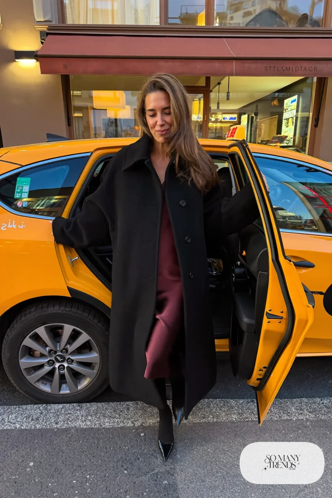 Woman in a long black cocoon coat over a wine satin midi slip dress, black opaque tights and black pointed pumps, outfit 5