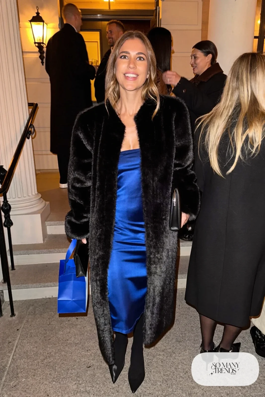 Woman in a black faux fur coat over a cobalt blue satin midi dress, opaque black tights and black pointed pumps, outfit 16