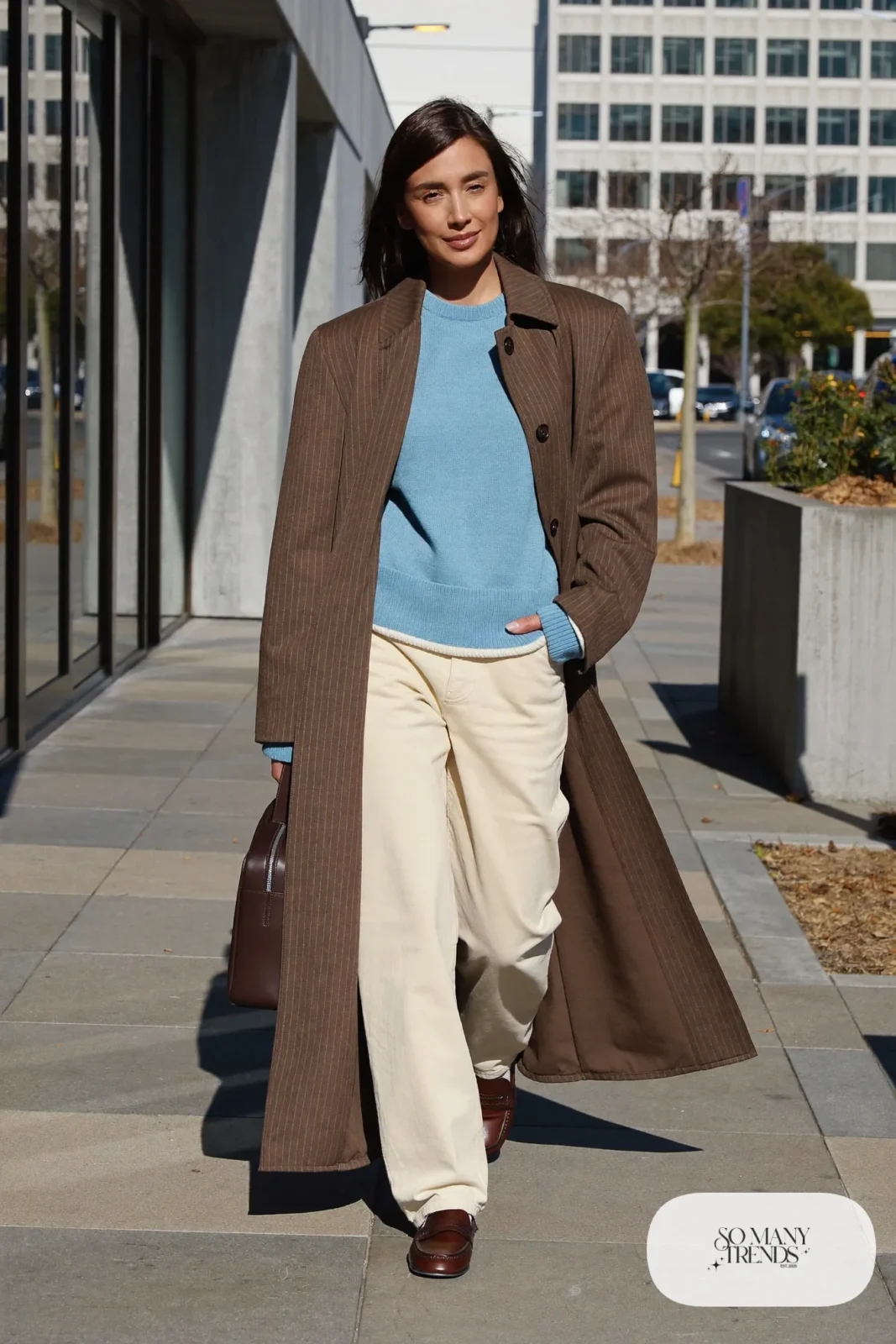 Woman in a long mocha pinstripe coat over a sky blue knit and cream pants with loafers and a bag, outfit 30