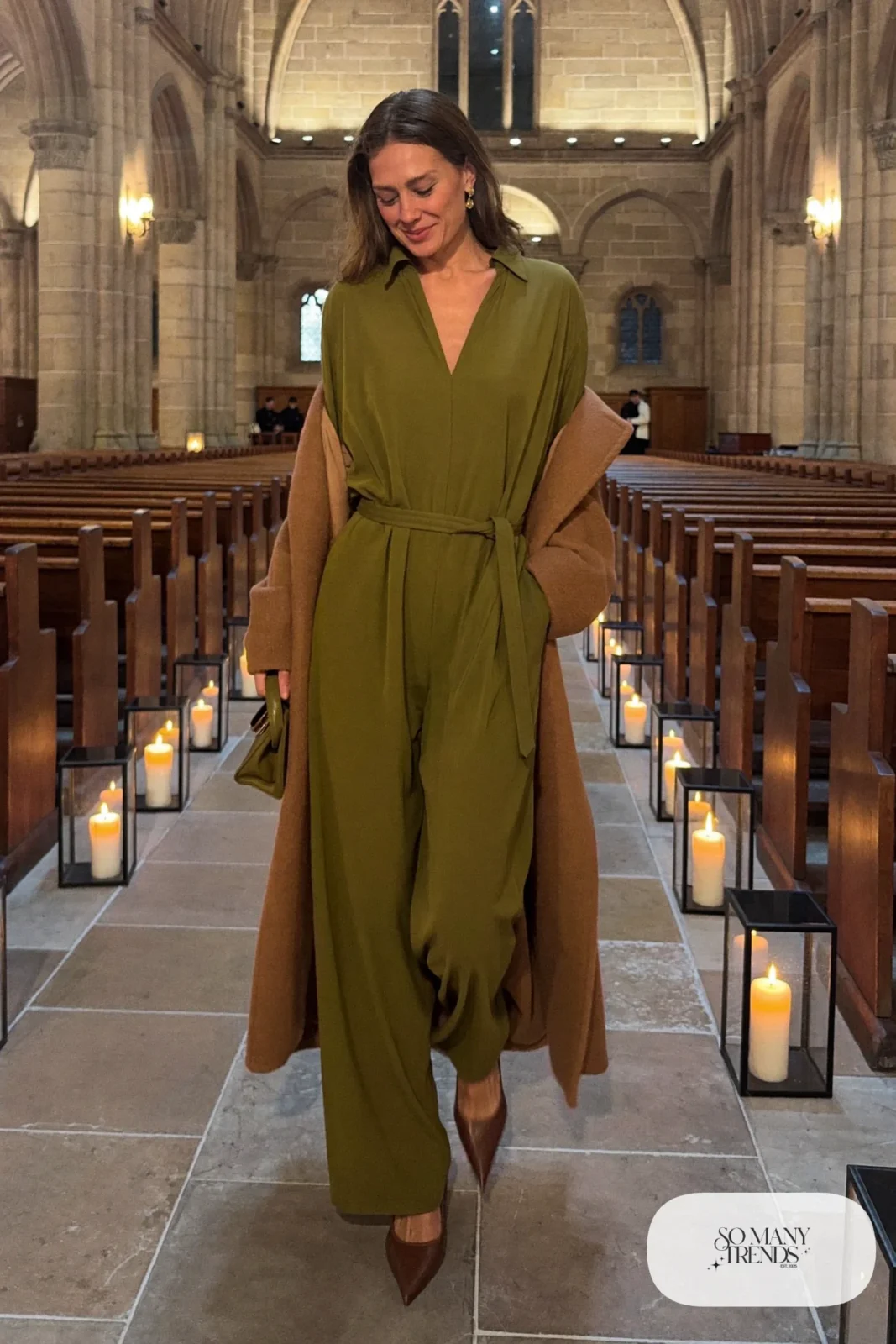 Woman in a moss green long sleeve wide-leg jumpsuit, long camel coat, cognac pointed heels and gold earrings, outfit 16