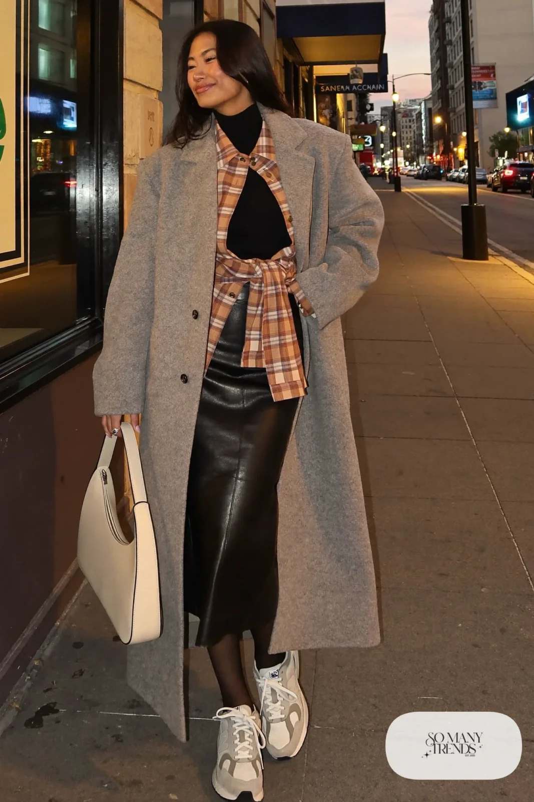 Woman in plaid flannel shirt tied at the waist, black faux-leather midi skirt, long gray coat and gray New Balance 9060 sneakers, outfit 12