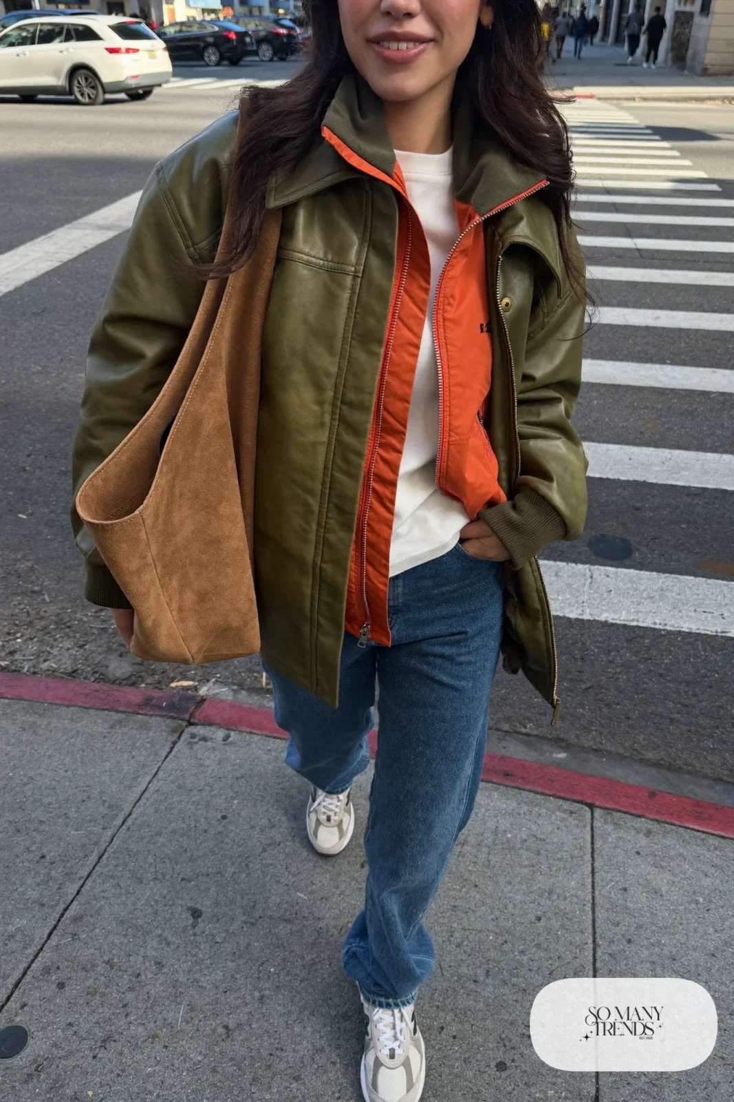 Woman in olive leather jacket over orange windbreaker, straight mid-wash jeans and gray New Balance 9060 sneakers, outfit 20