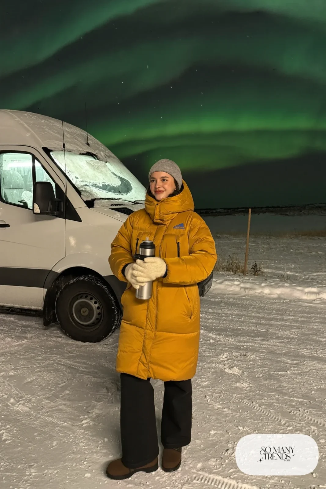 Woman in a mustard down parka, black straight pants, brown insulated lug-sole boots and a gray knit beanie under a night sky with northern lights, outfit 1