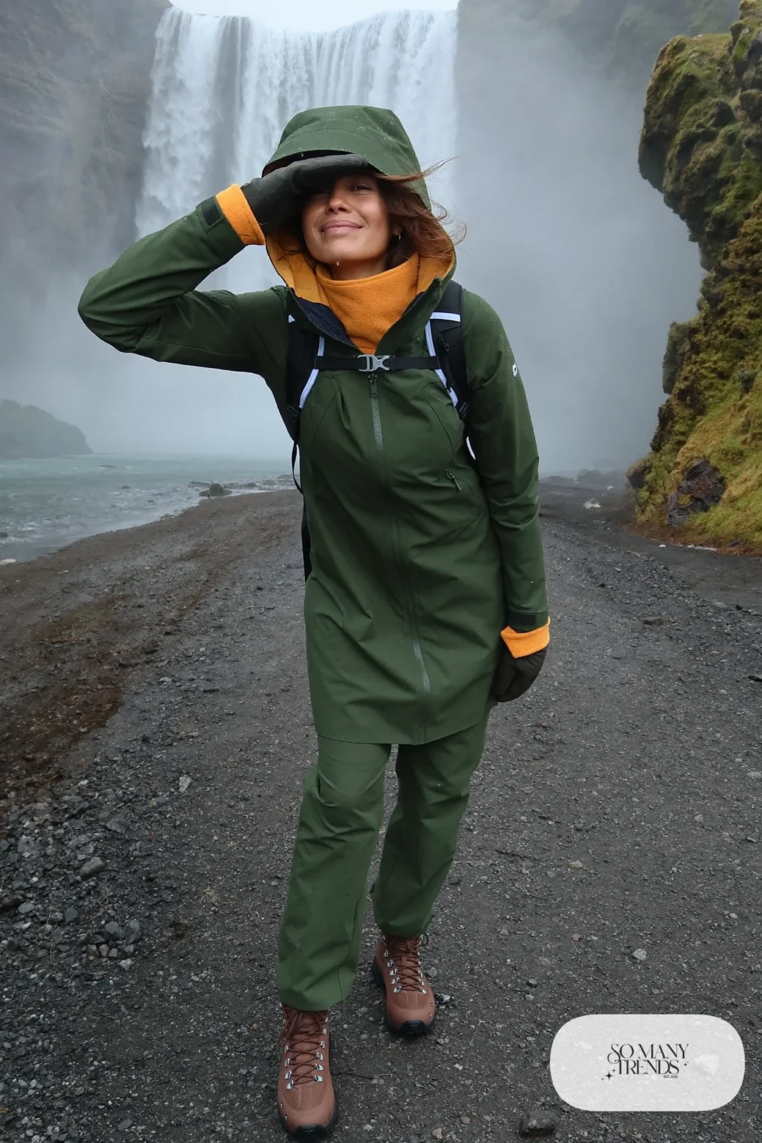 Woman in a green waterproof rain jacket and pants with brown waterproof boots in front of a waterfall in winter, outfit 8