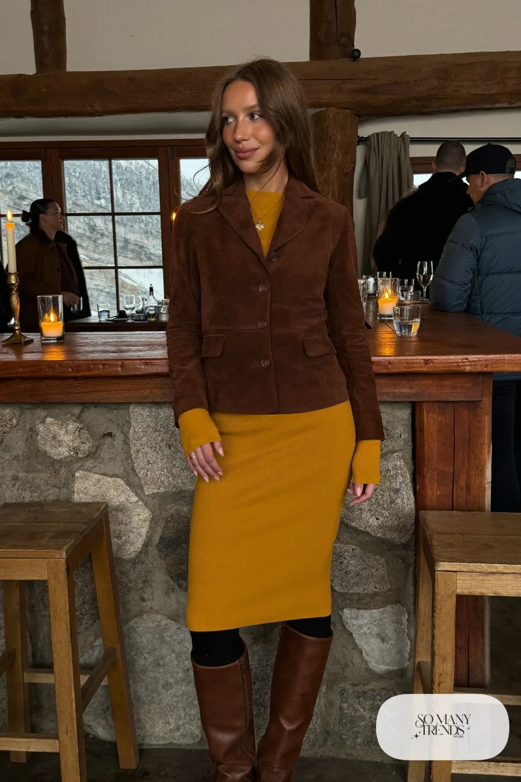 Woman in a chocolate suede jacket, mustard knit midi dress, black tights and tall brown boots at a lodge bar, outfit 18
