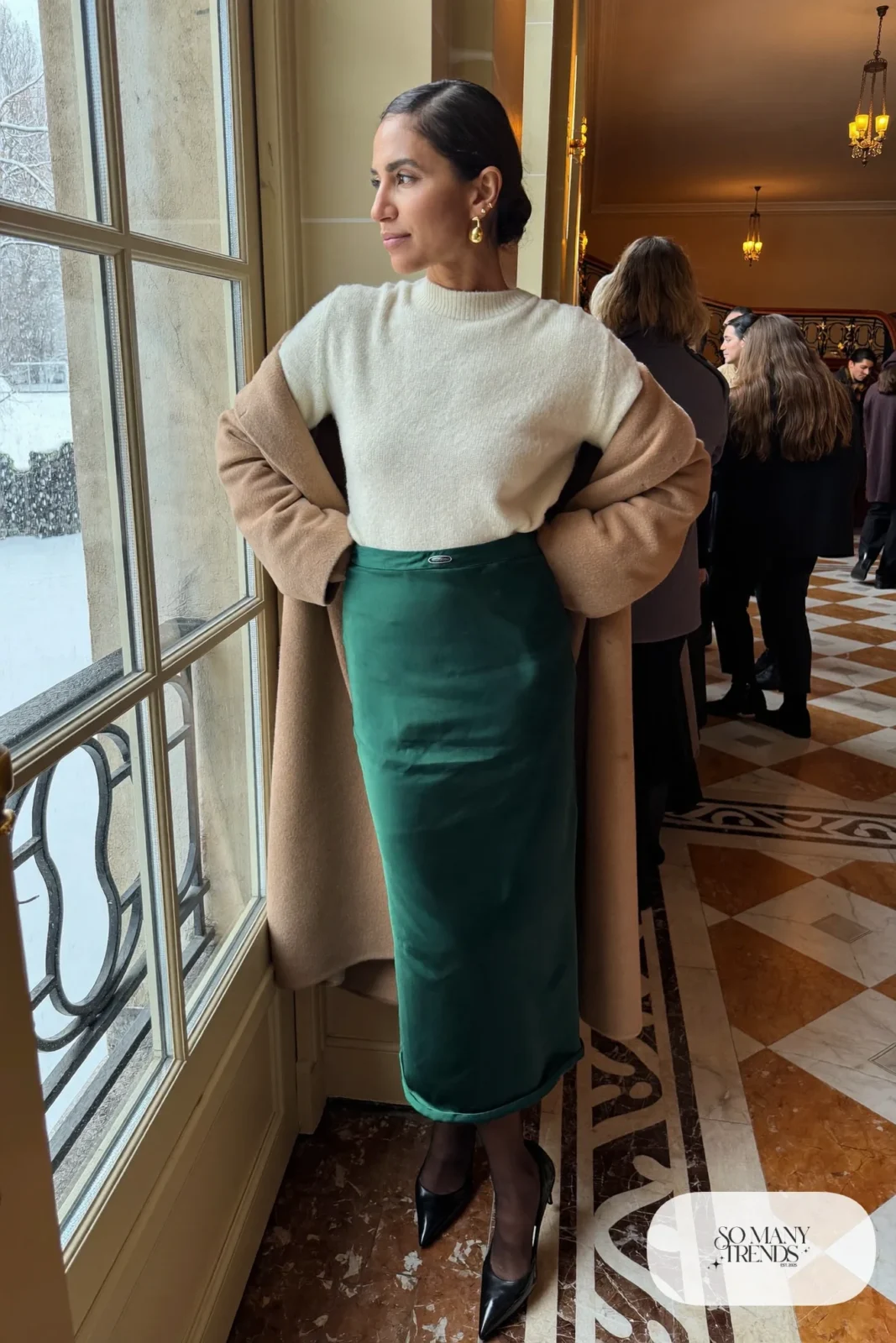 Woman in a forest green satin midi skirt, cream cashmere sweater, long camel coat, sheer tights and black pumps, theater outfit 24