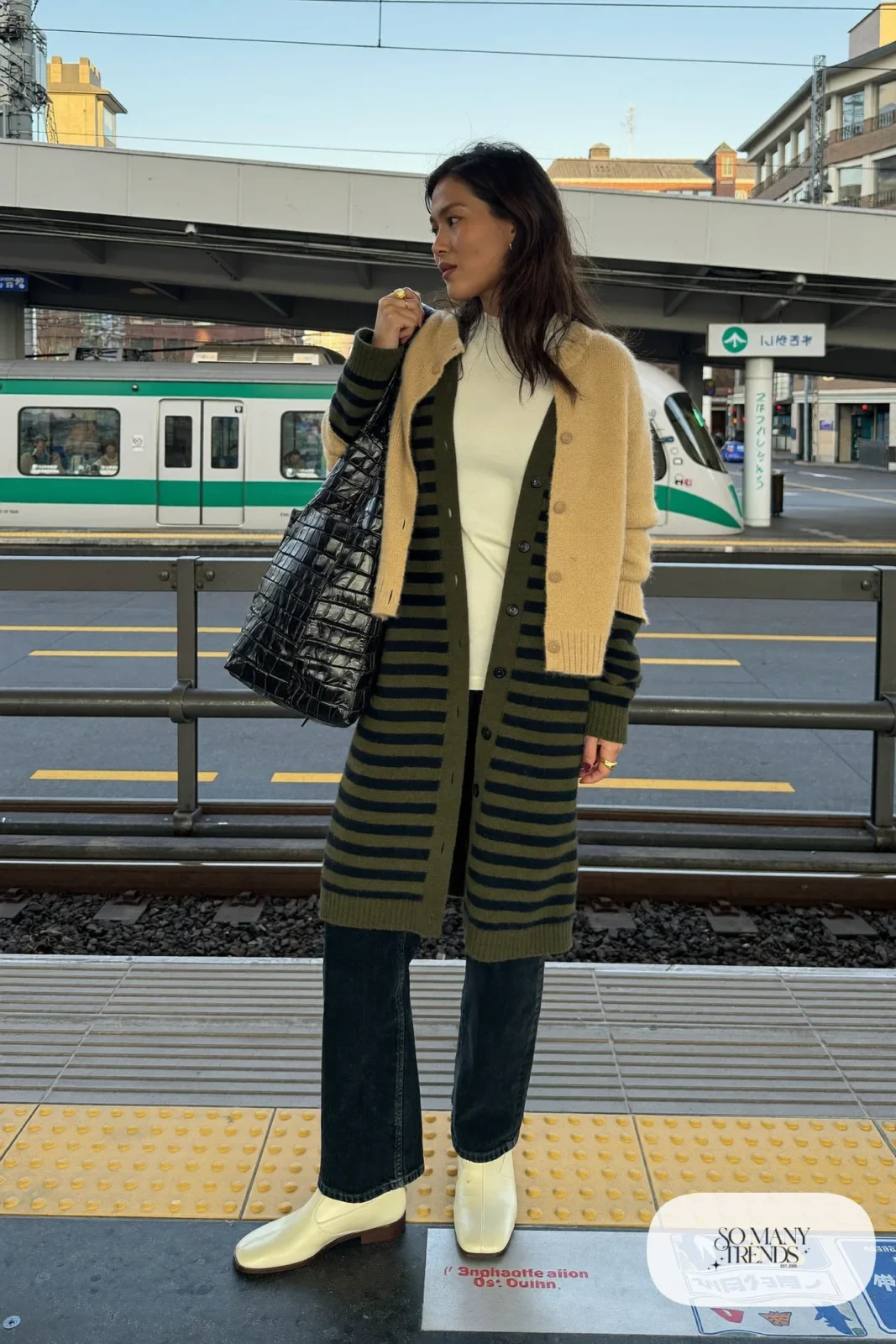 Woman in a sand knit cardigan jacket over a long olive striped cardigan, white tee, black straight jeans and cream flat ankle boots with a black tote, outfit 9