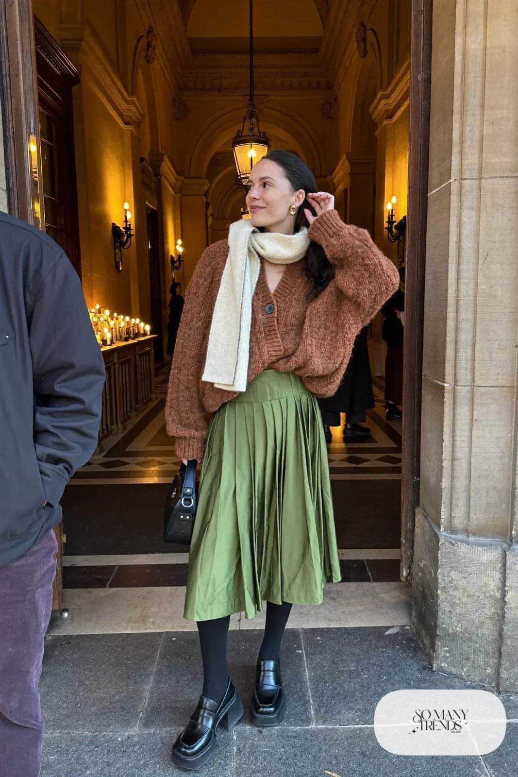 Woman in a moss green pleated satin midi skirt, cognac chunky knit sweater and black heeled loafers, outfit 9