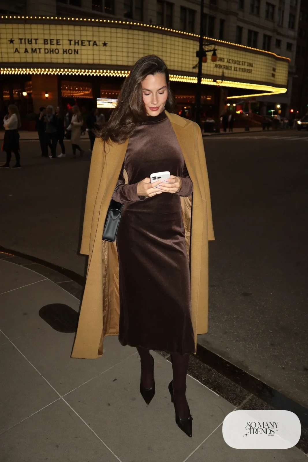 Woman in a chocolate brown velvet midi dress, camel wool overcoat, dark tights and black pumps, theater outfit 3