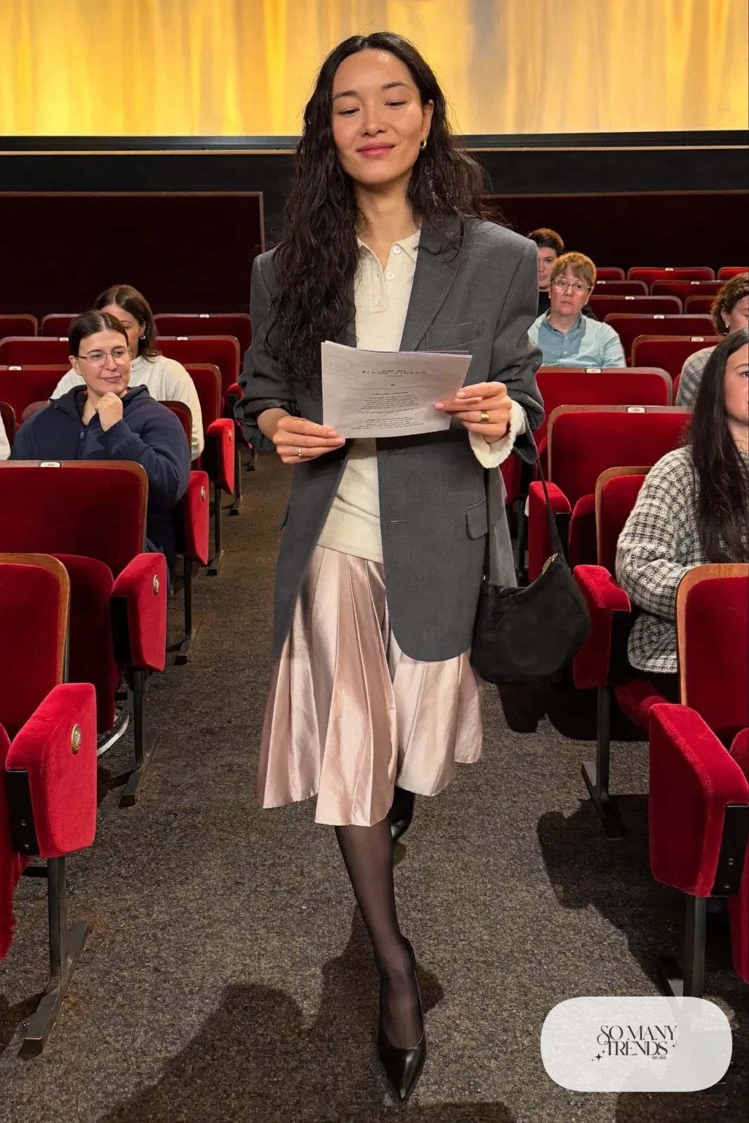 Woman in a dusty pink pleated satin midi skirt, charcoal oversized blazer, cream knit top and black pointed pumps, theater outfit 8