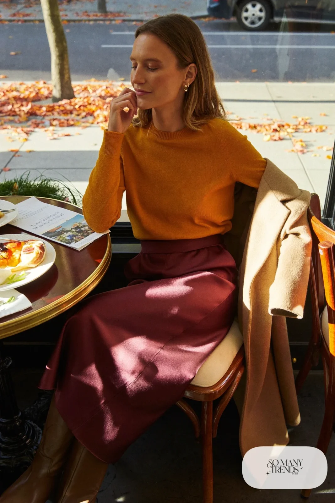 Woman in a mustard knit sweater, burgundy satin midi skirt, camel coat and brown knee-high boots, theater outfit 30