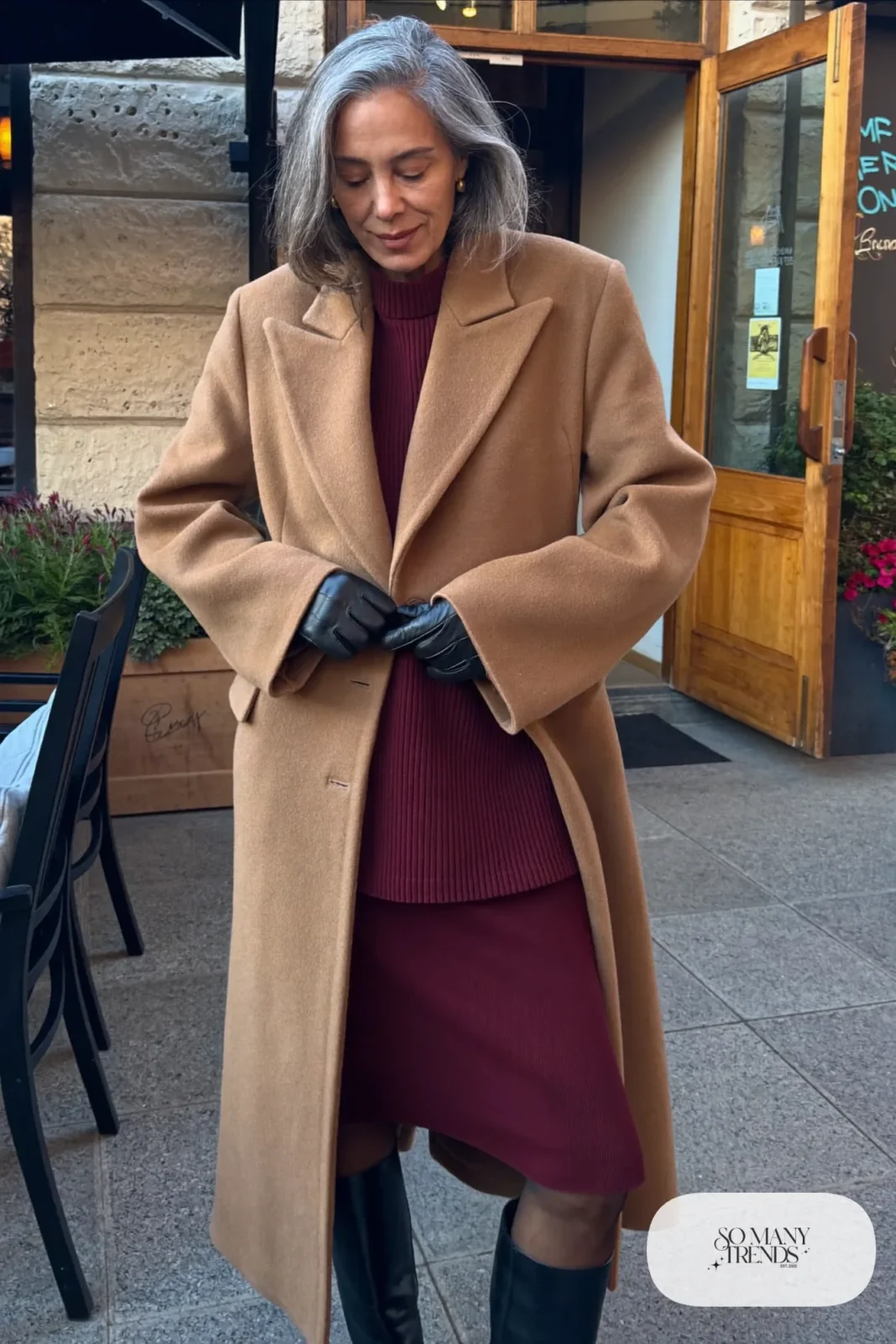 Woman in her sixties wearing a burgundy knit top and midi skirt set with a long camel coat and tall black boots, outfit 24