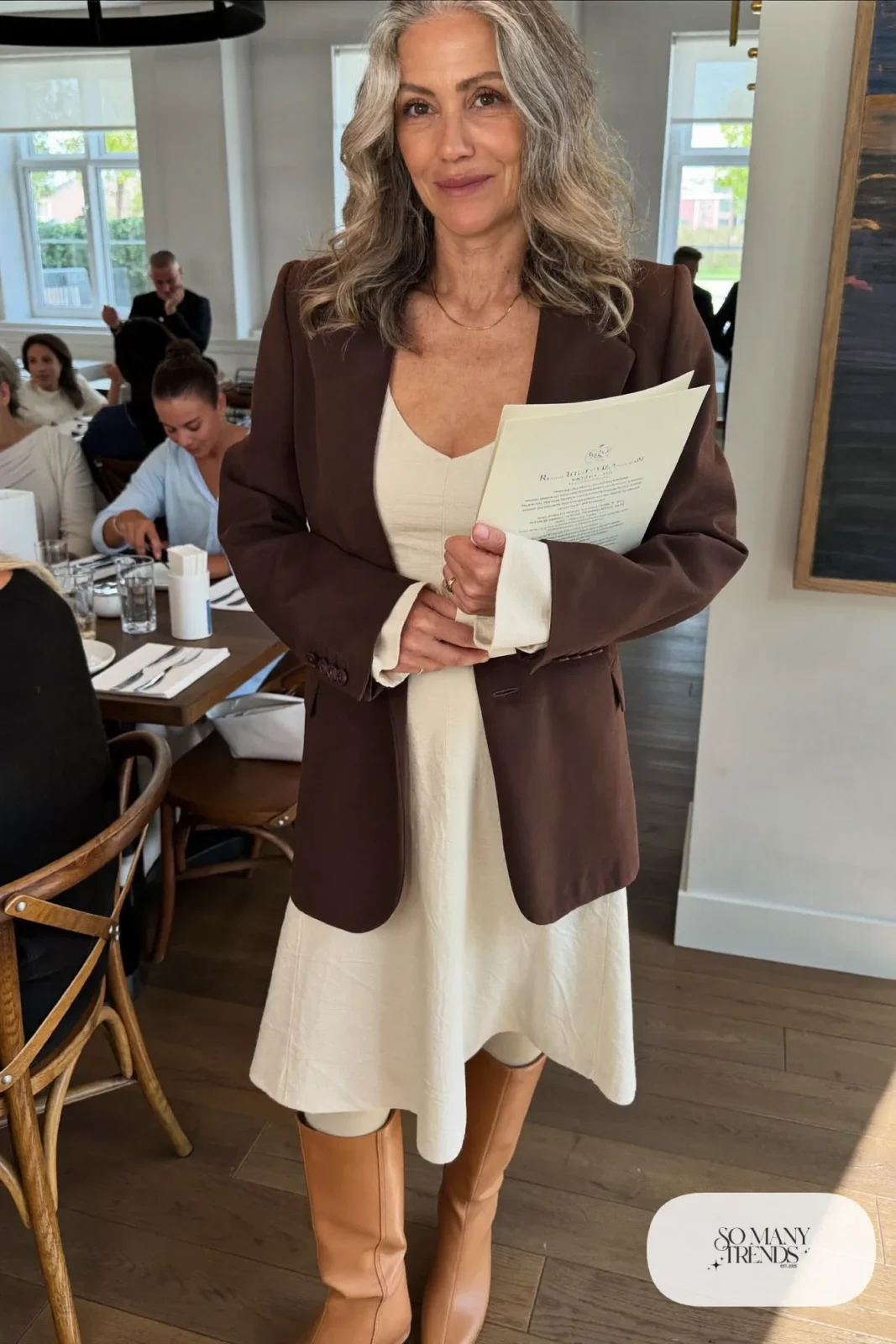 Woman in her sixties wearing a long chocolate blazer over a cream midi dress with tan boots, outfit 31